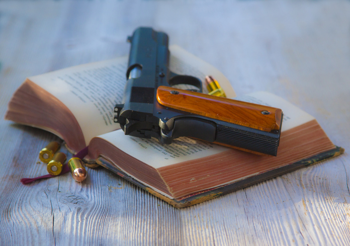 Handgun laying on top of a old open bible with bullets around it, on a rustic wood table
