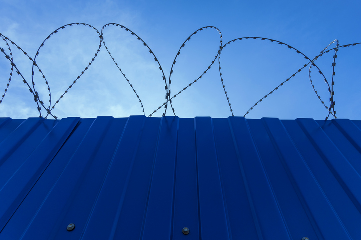 Solid, blue iron fence with barbed wire under a blue sky.