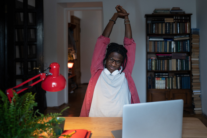 Tired sleepy African American man freelancer pulls arms up to stretch after difficult workimg day