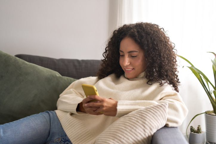 Latin young woman sitting on couch using mobile phone at home.