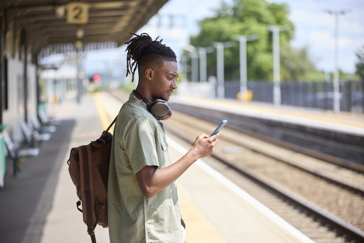 Young Man Commuting To Work On Train Standing On Platform Looking At Mobile Phone For Travel Information Or Social Media