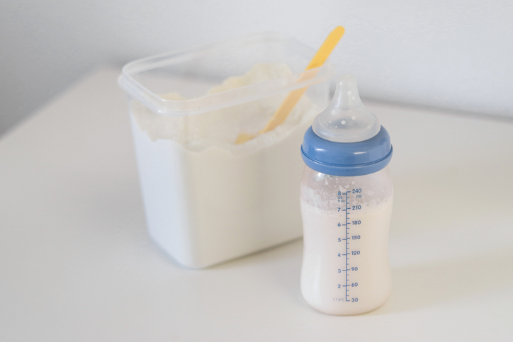 A feeding bottle with baby formula milk on white drawer, dry powder milk in a can with plastic scoop in background