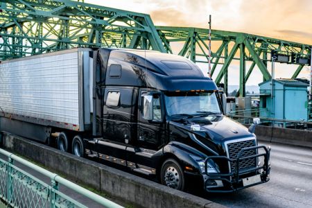 Black shiny big rig semi truck transporting frozen cargo in refrigerator semi trailer running on the truss arched bridge at twilight time