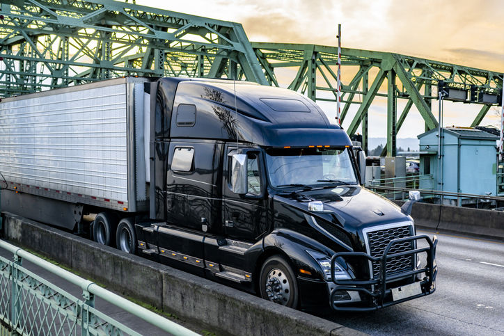 Black shiny big rig semi truck transporting frozen cargo in refrigerator semi trailer running on the truss arched bridge at twilight time