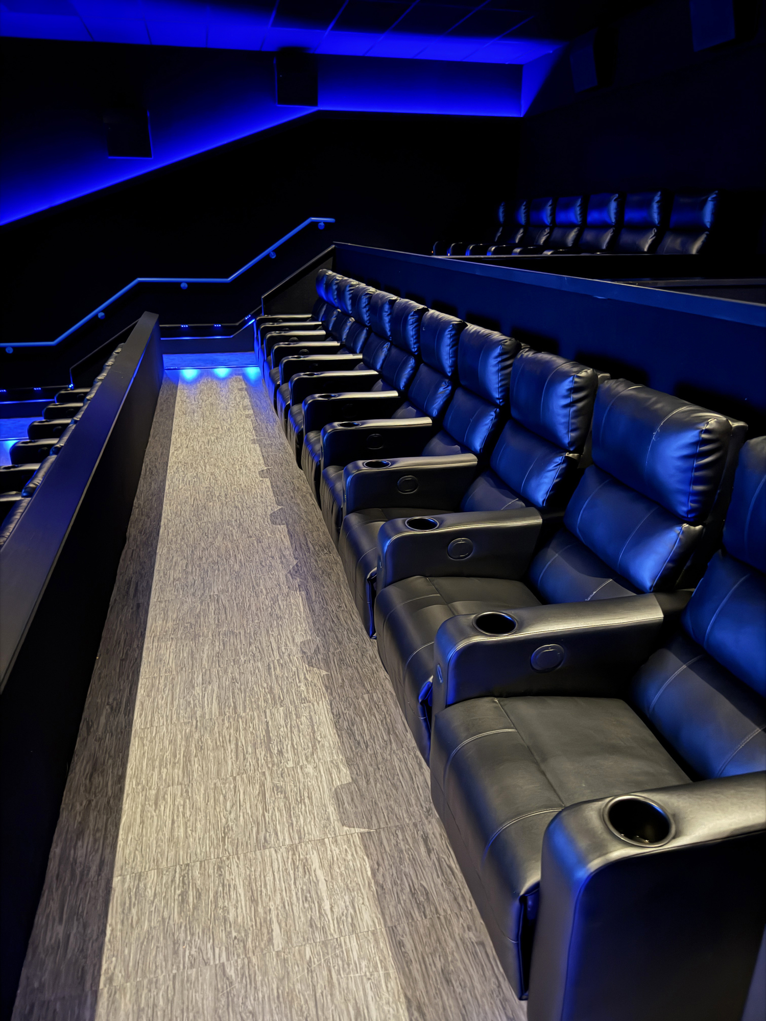 Image of empty cinema, rows of black leather chair seats, tiered armed seating with cupholders in armrests, ambient lighting, focus on foreground