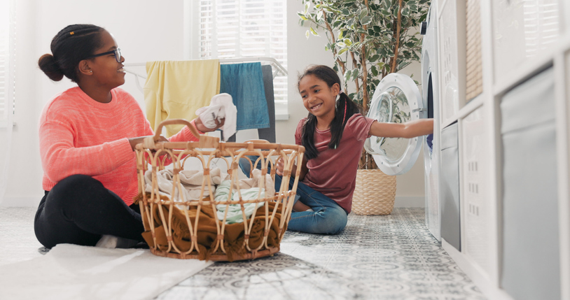 A mother sits on the bathroom floor, organizing laundry. She passes bright clothes to her school-aged daughter who helps by tossing them one by one into the washing machine.