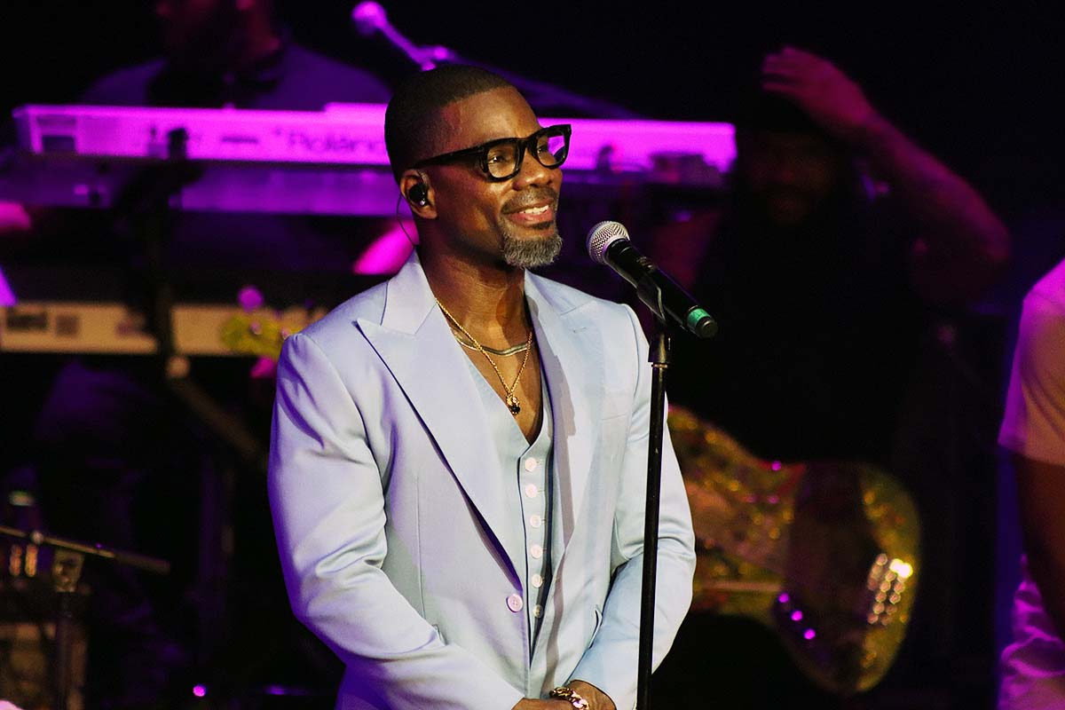 A man with a beard and glasses speaking into a microphone on a stage with purple lighting.