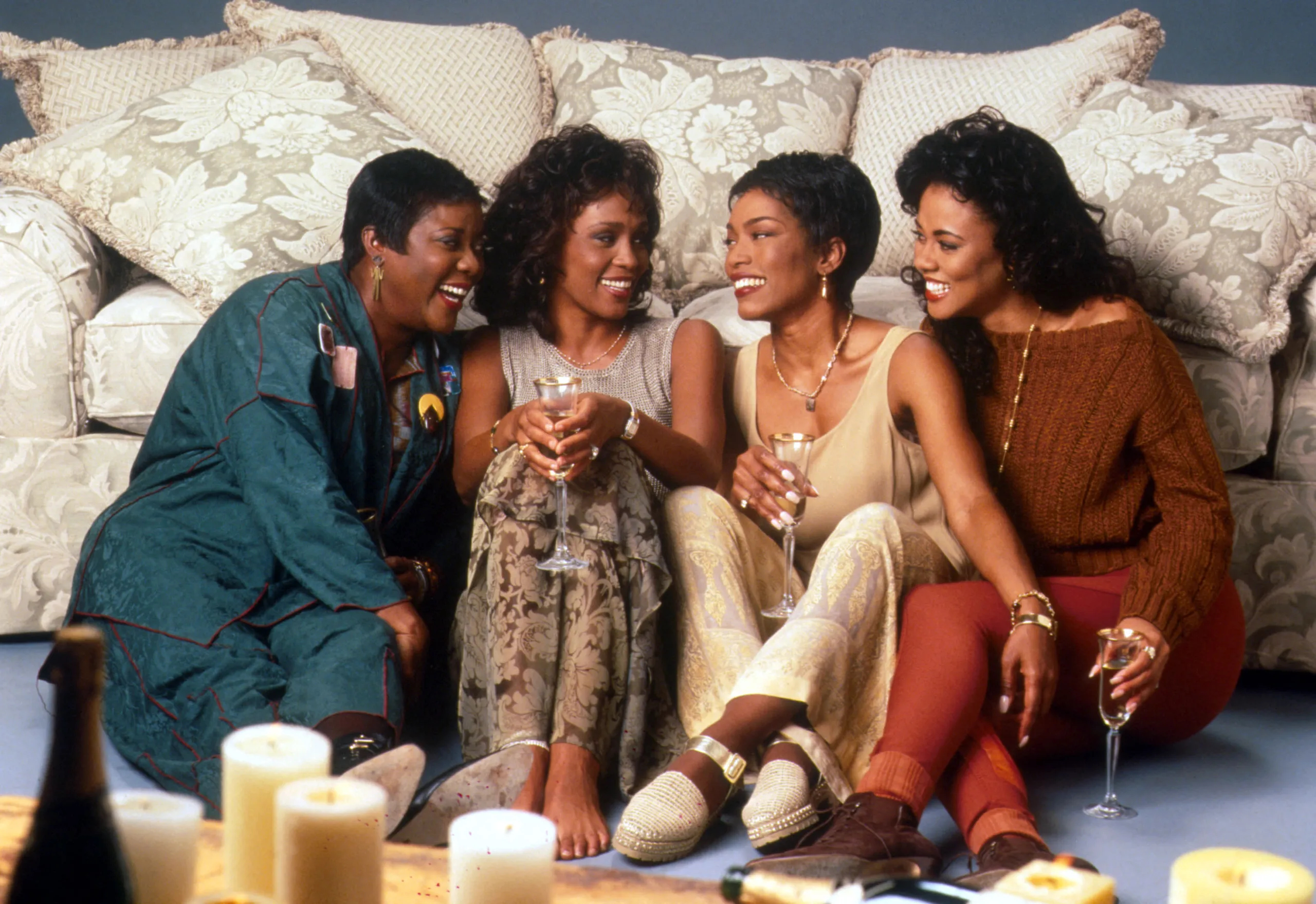 Four women sitting on a couch, smiling and holding drinks, in a cozy living room setting.