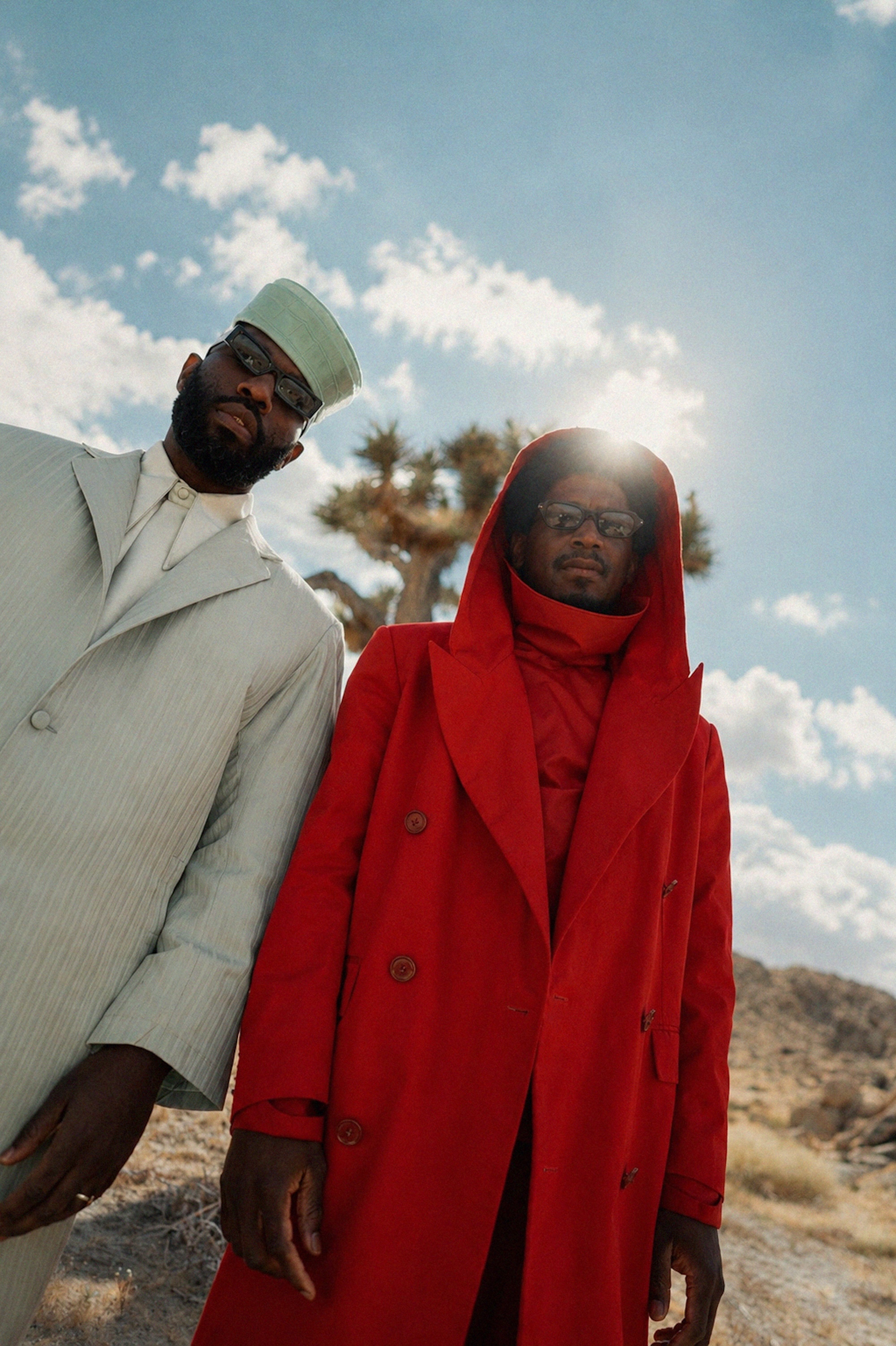 Two people wearing distinctive outerwear - a man in a gray coat and a woman in a bright red coat - standing in a desert landscape with a cloudy sky.