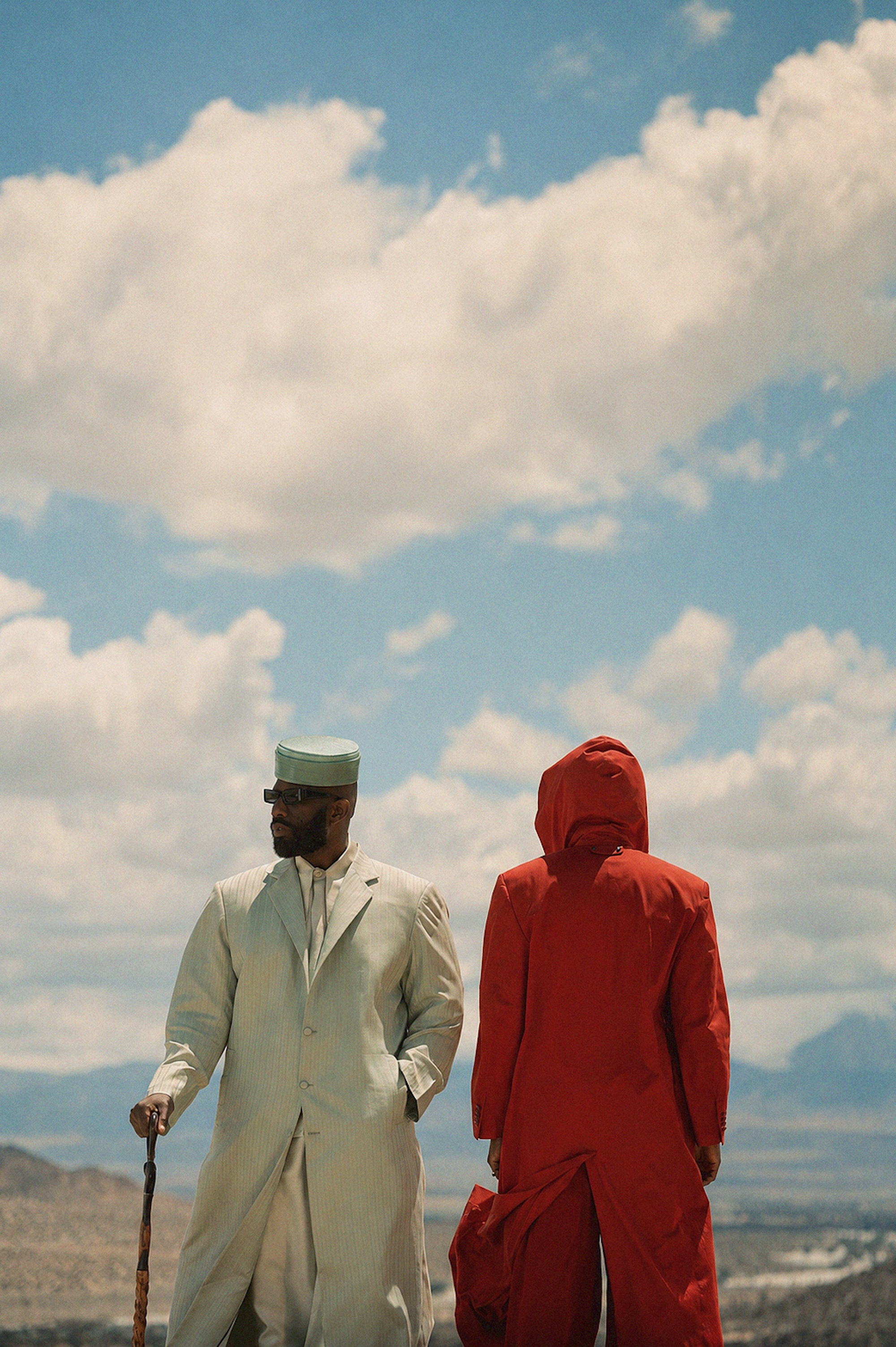 Two people, one in a white robe and one in a red hooded robe, walking against a cloudy blue sky.