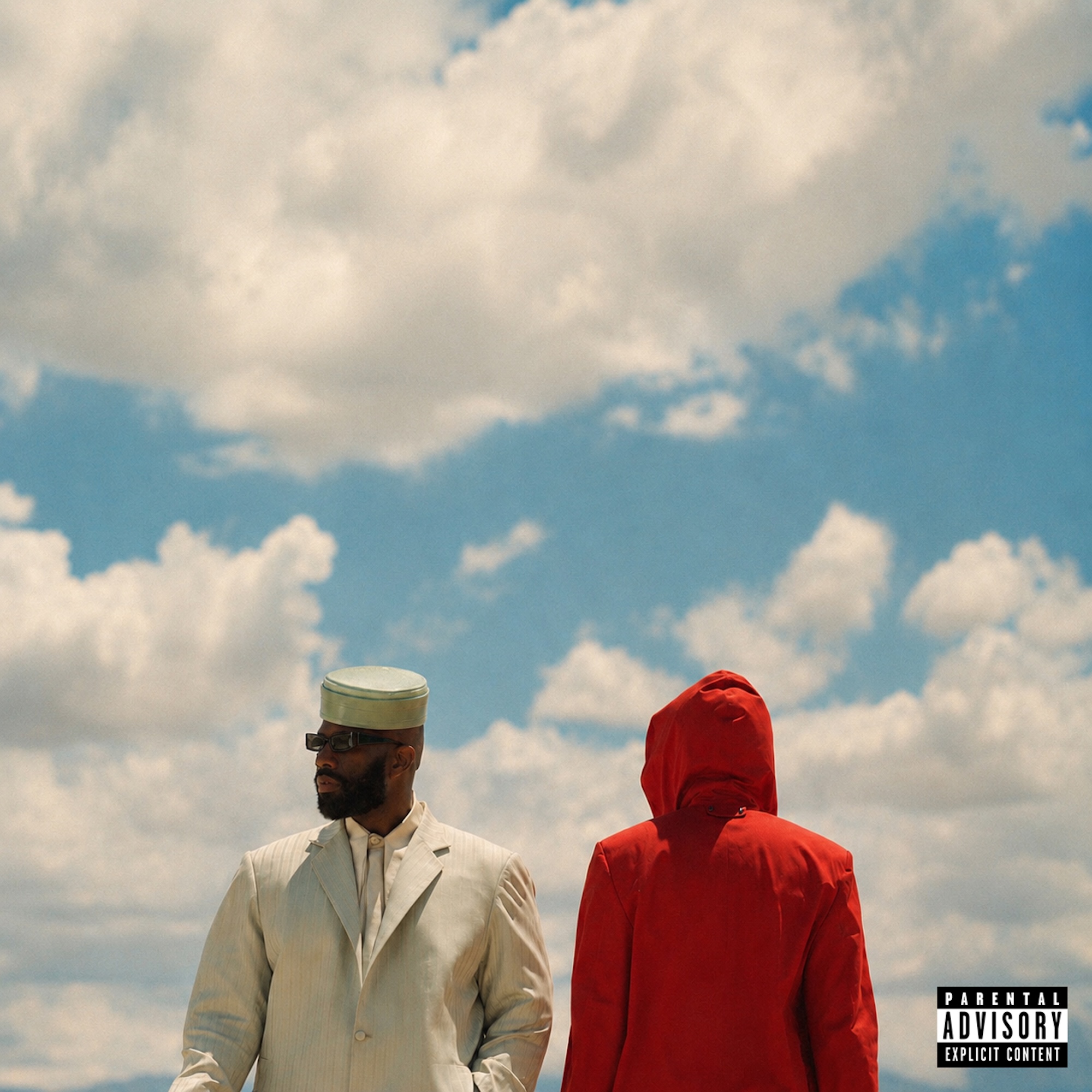 Two figures, one in a white suit and one in a red hooded jacket, stand against a cloudy sky.