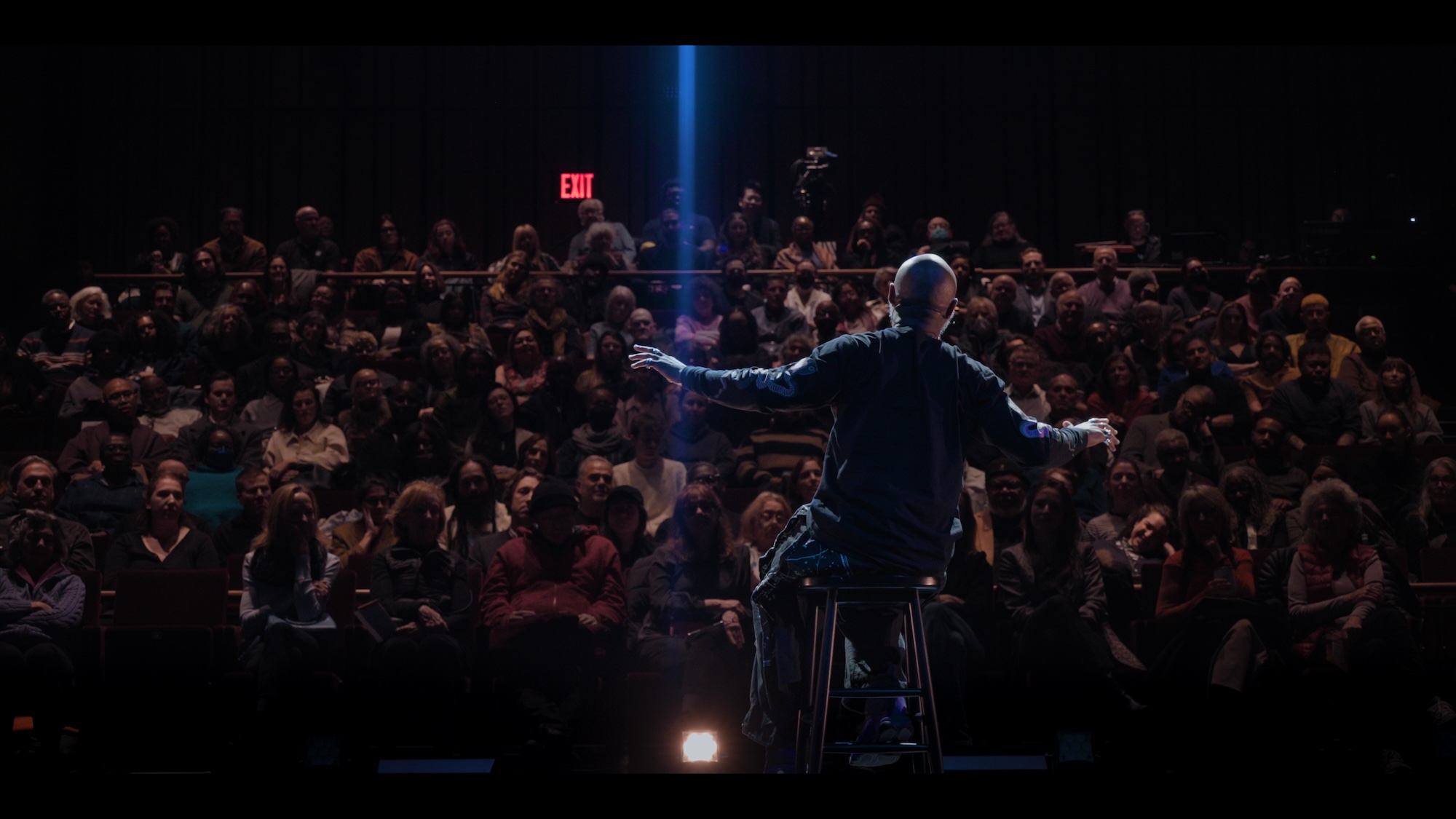A large crowd of people in a dark auditorium, with a single person standing on a stage and gesturing to the audience.