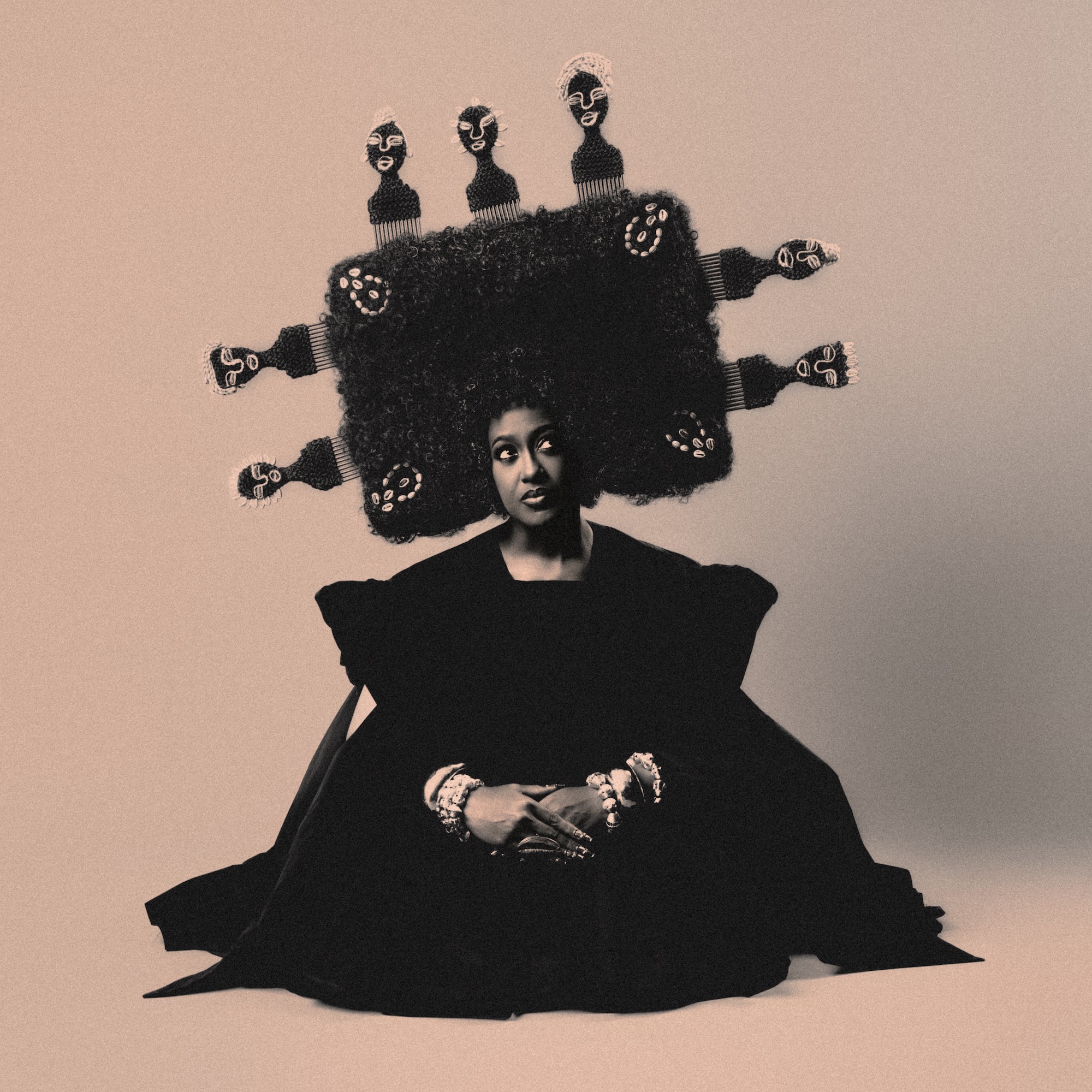 A person with an elaborate, sculptural black hairstyle wearing a large black robe and multiple bracelets, sitting against a beige background.