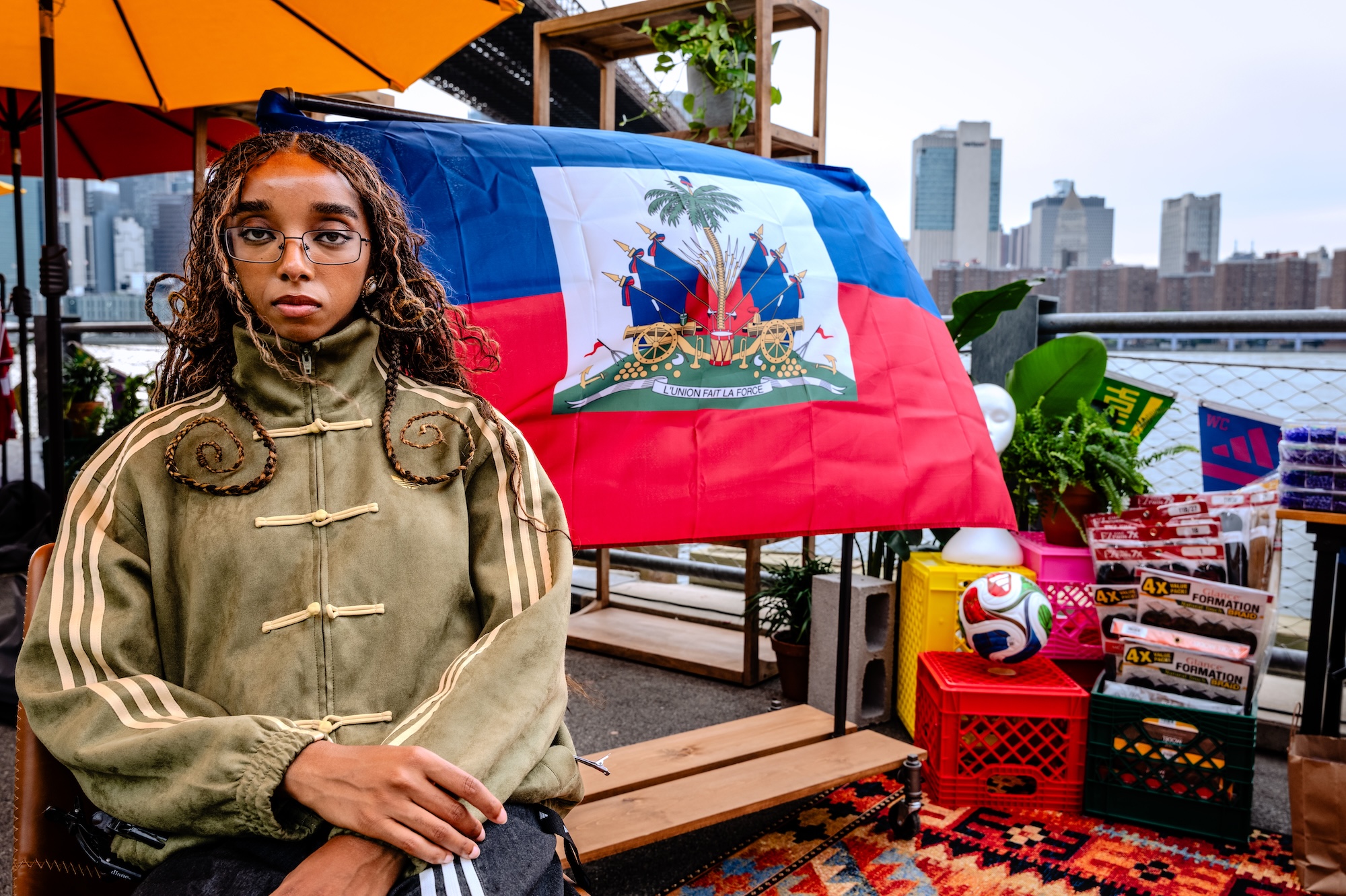 A person wearing a green jacket and glasses stands in front of a Haitian flag backdrop, surrounded by various items including a soccer ball.