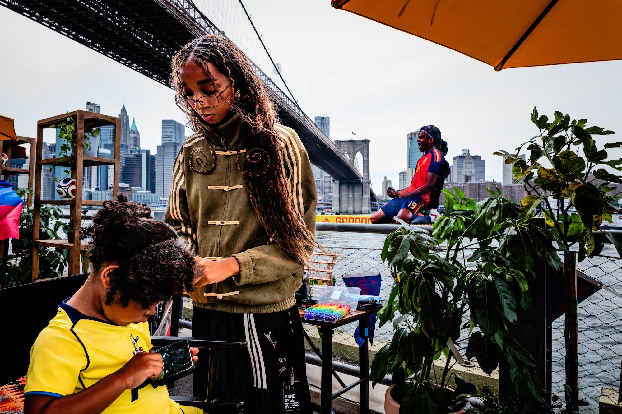 A person in a green jacket stands in front of the Brooklyn Bridge, with a person in a red shirt sitting on a bench behind them, and plants and umbrellas in the foreground.