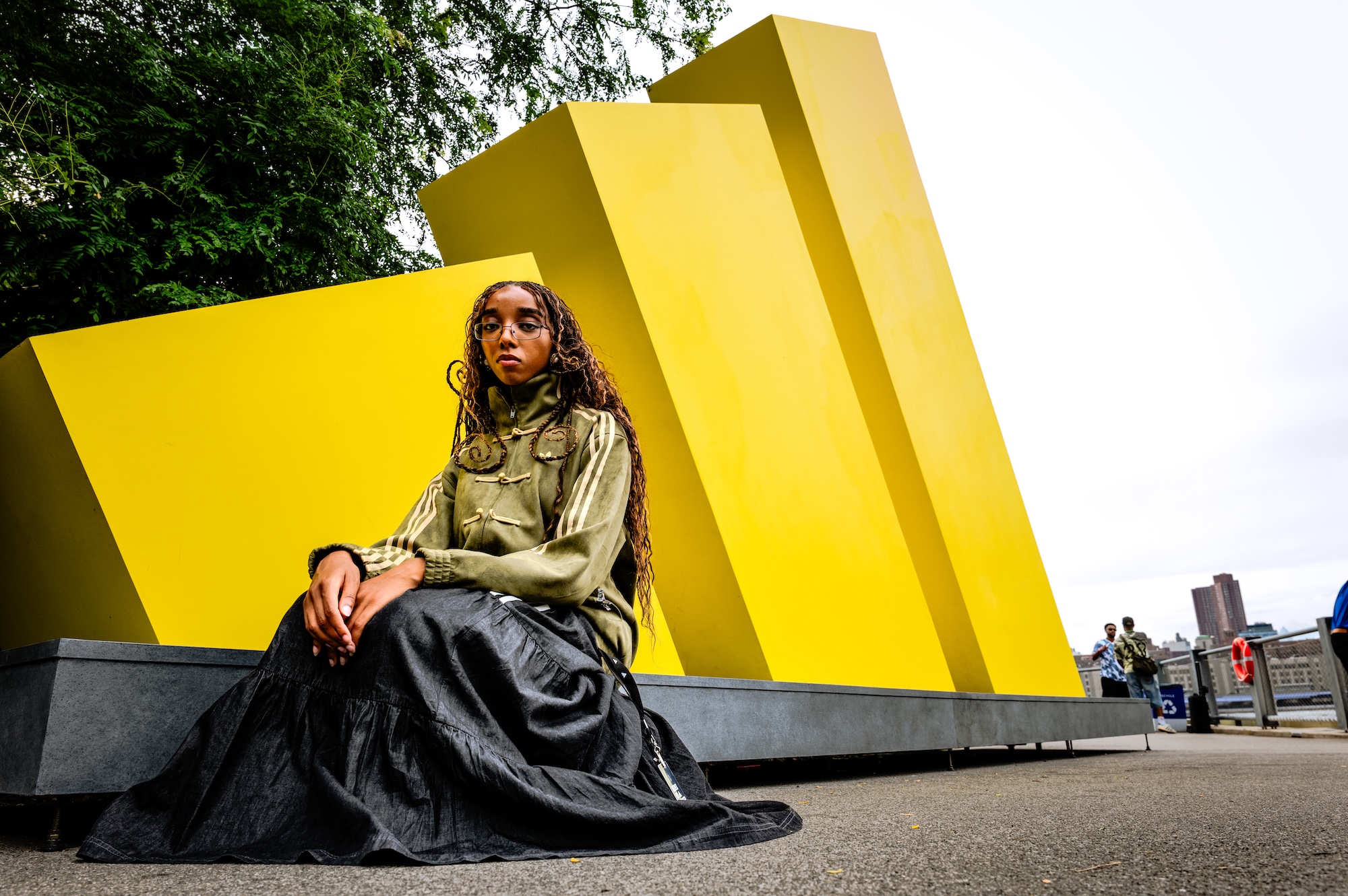 A person wearing a green military-style jacket and black skirt sits on a platform in front of large yellow geometric structures in an urban setting.