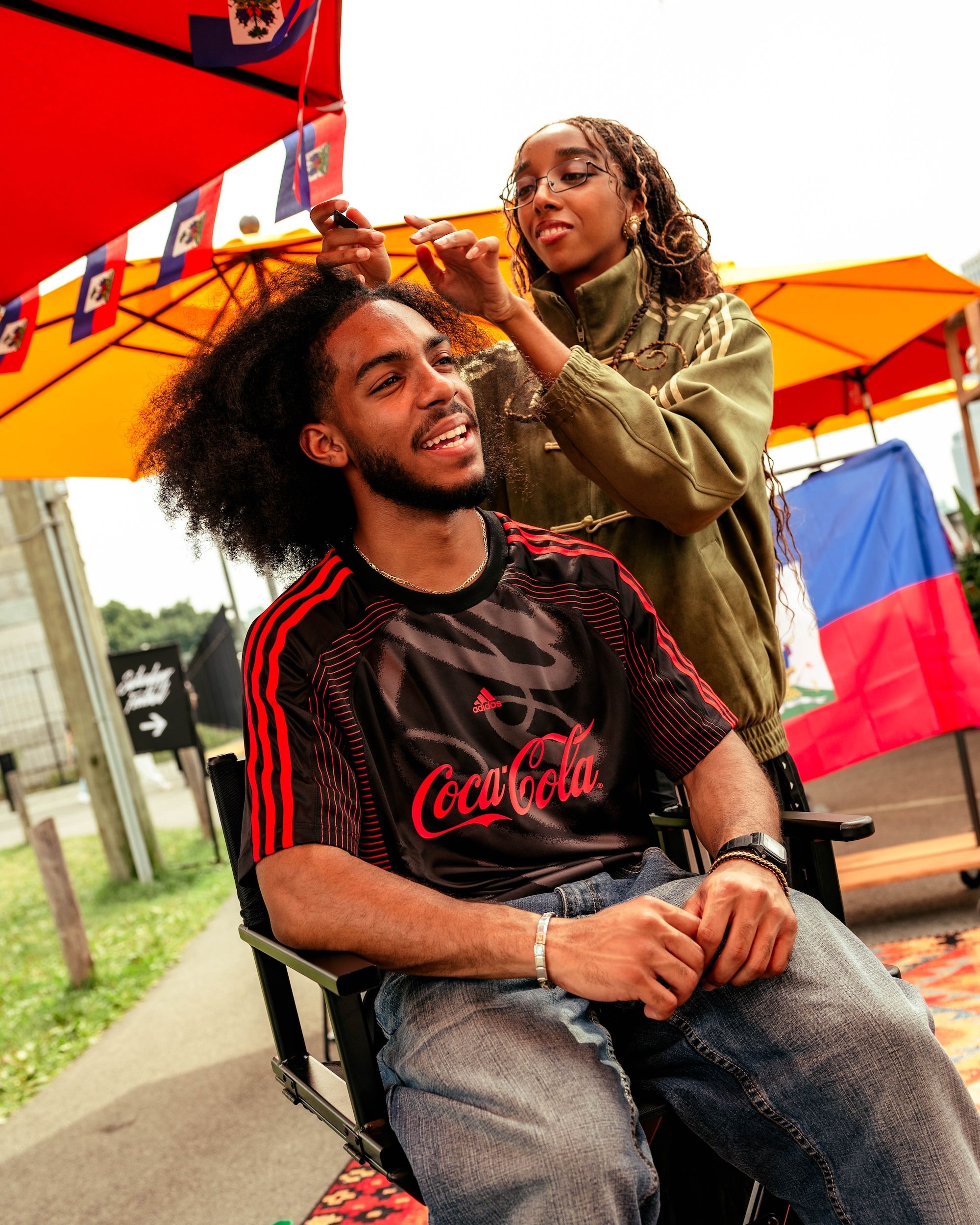 A person in a Coca-Cola jersey smiles as another person styles their hair in an outdoor setting with colorful umbrellas and flags.