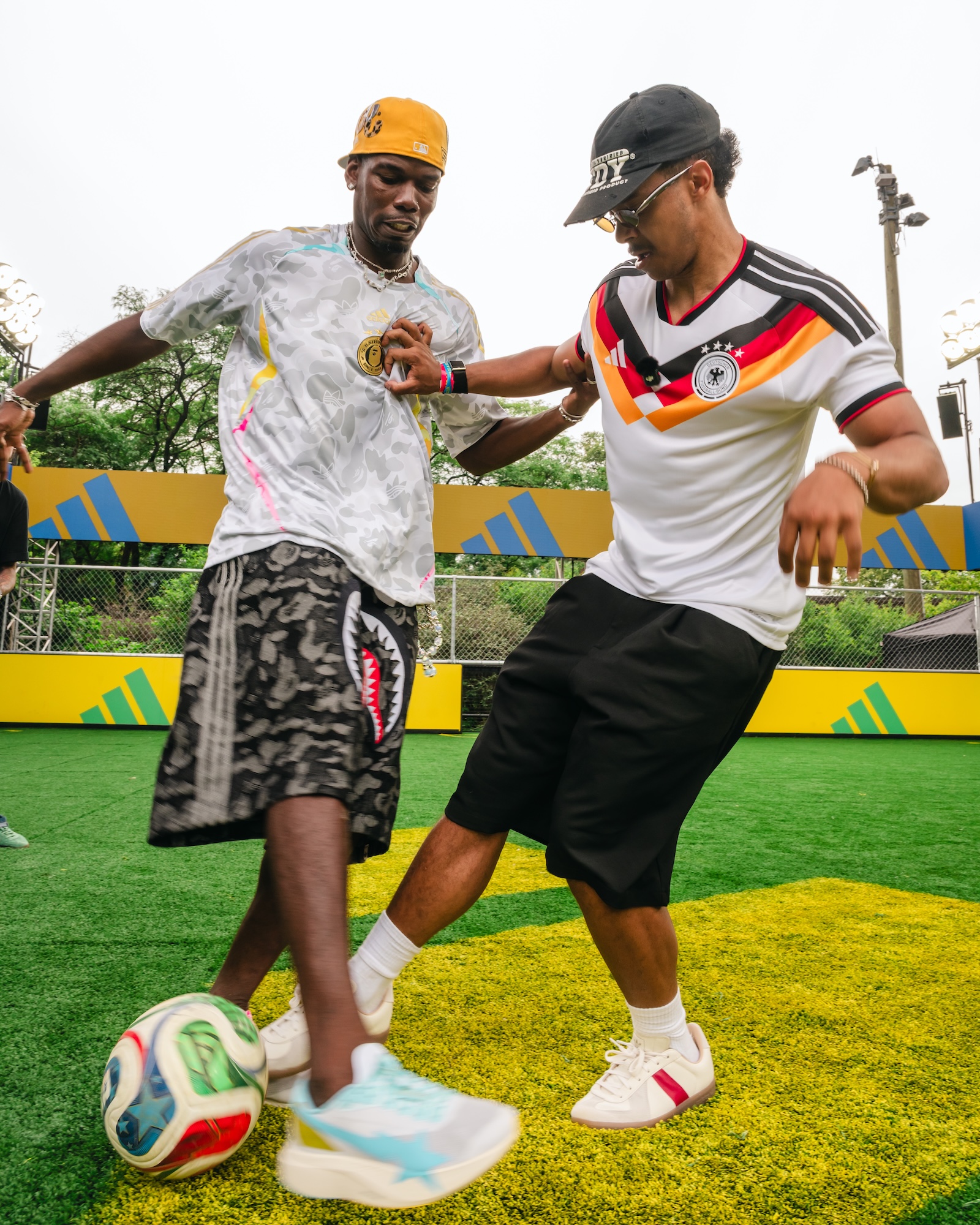Two young men wearing sports jerseys and hats, one in a white jersey with the German national team logo, the other in a black jersey with the German national team logo, engaged in a friendly interaction on a soccer field.