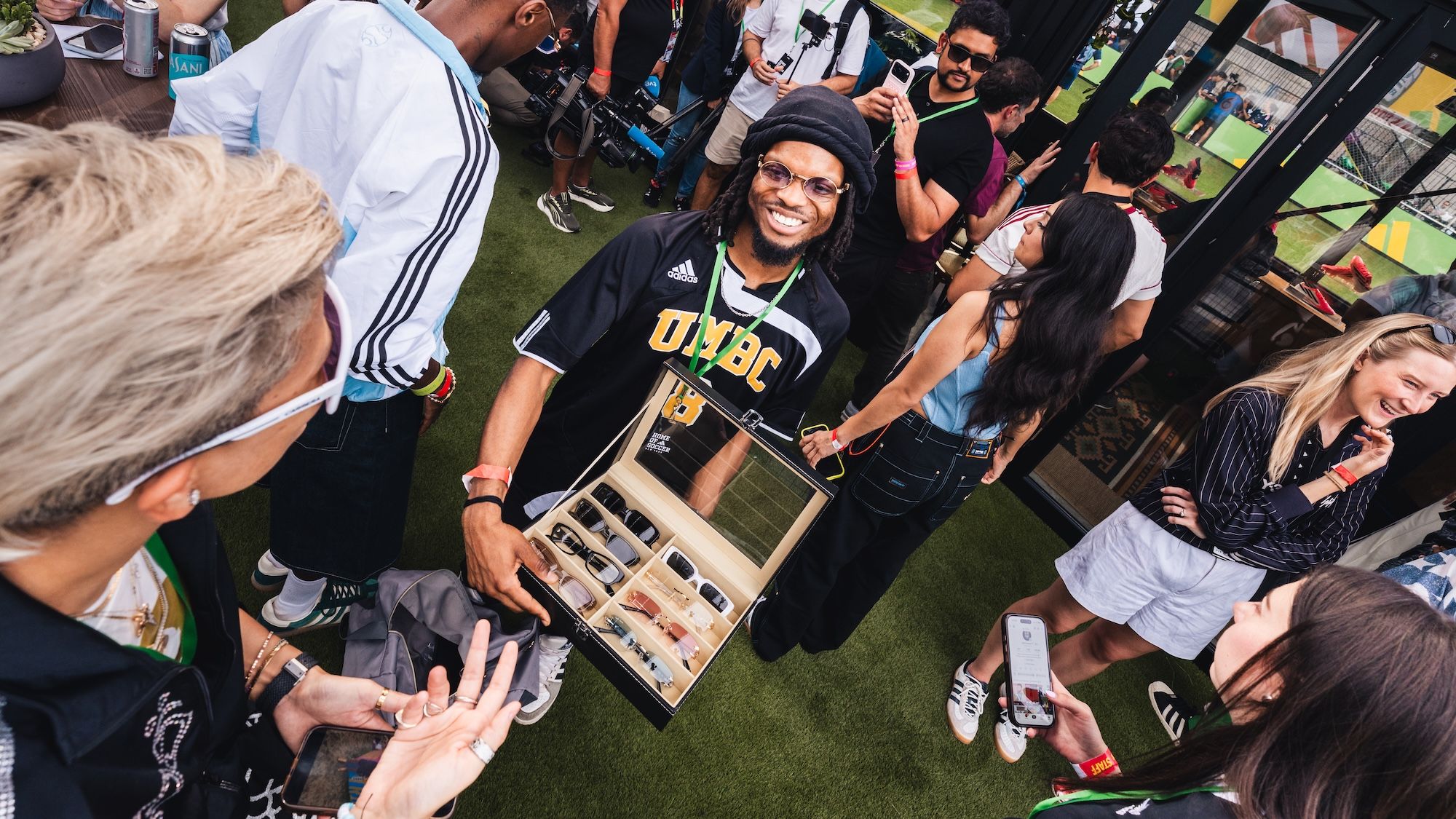 A group of people gathered around a person holding a box of merchandise, smiling and interacting with each other. The person is wearing a black shirt with "UNBC" printed on it.