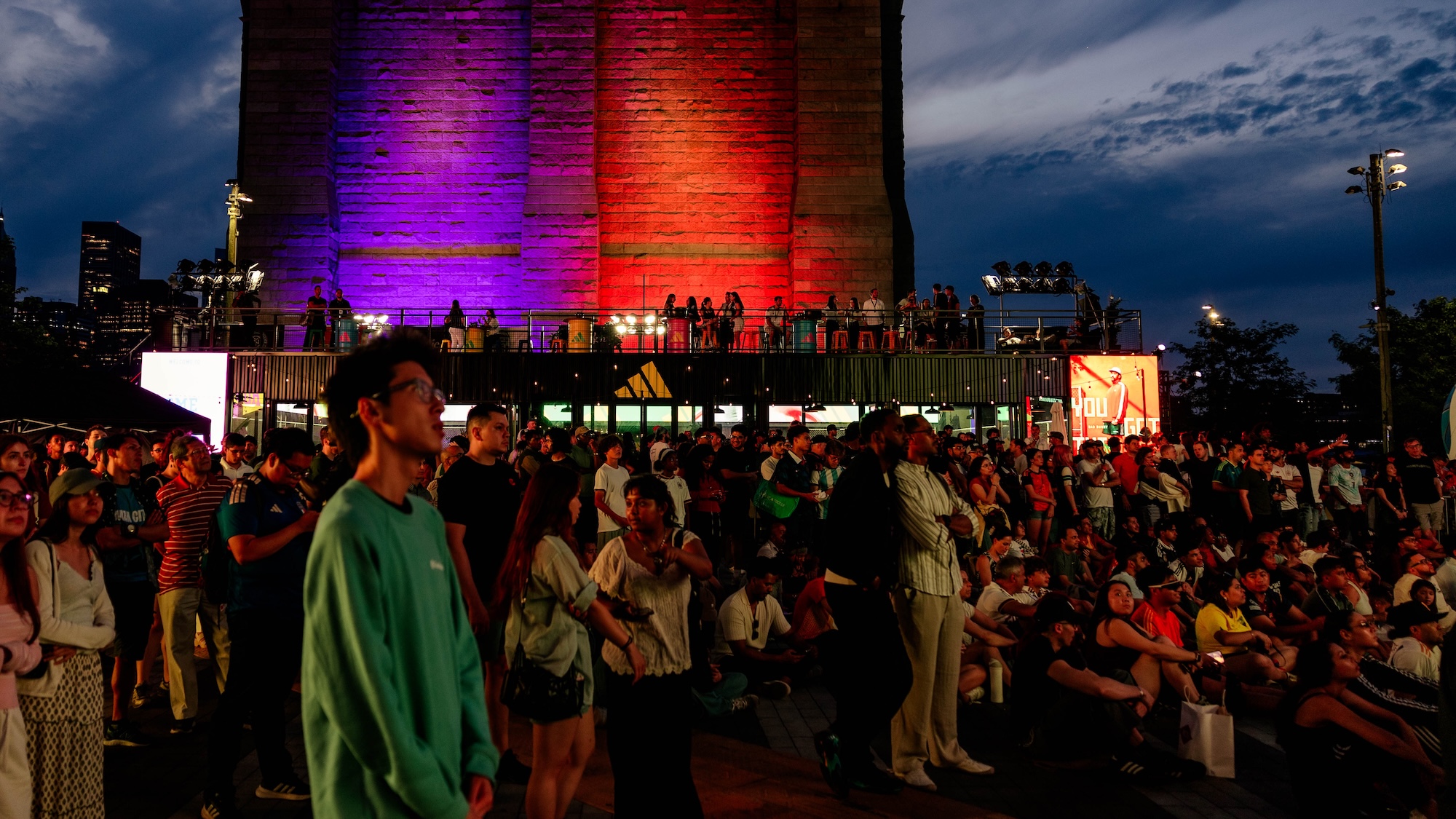Nighttime crowd gathered in front of a brightly lit building with purple and orange lights, people standing and sitting on a raised platform.
