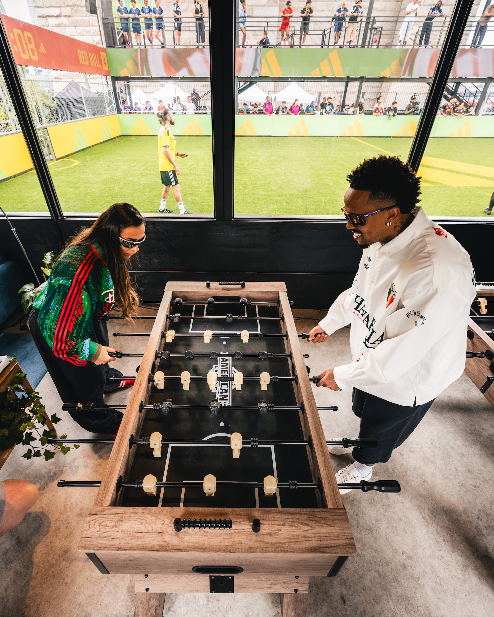 Two people playing foosball in a sports facility with spectators visible through the window.