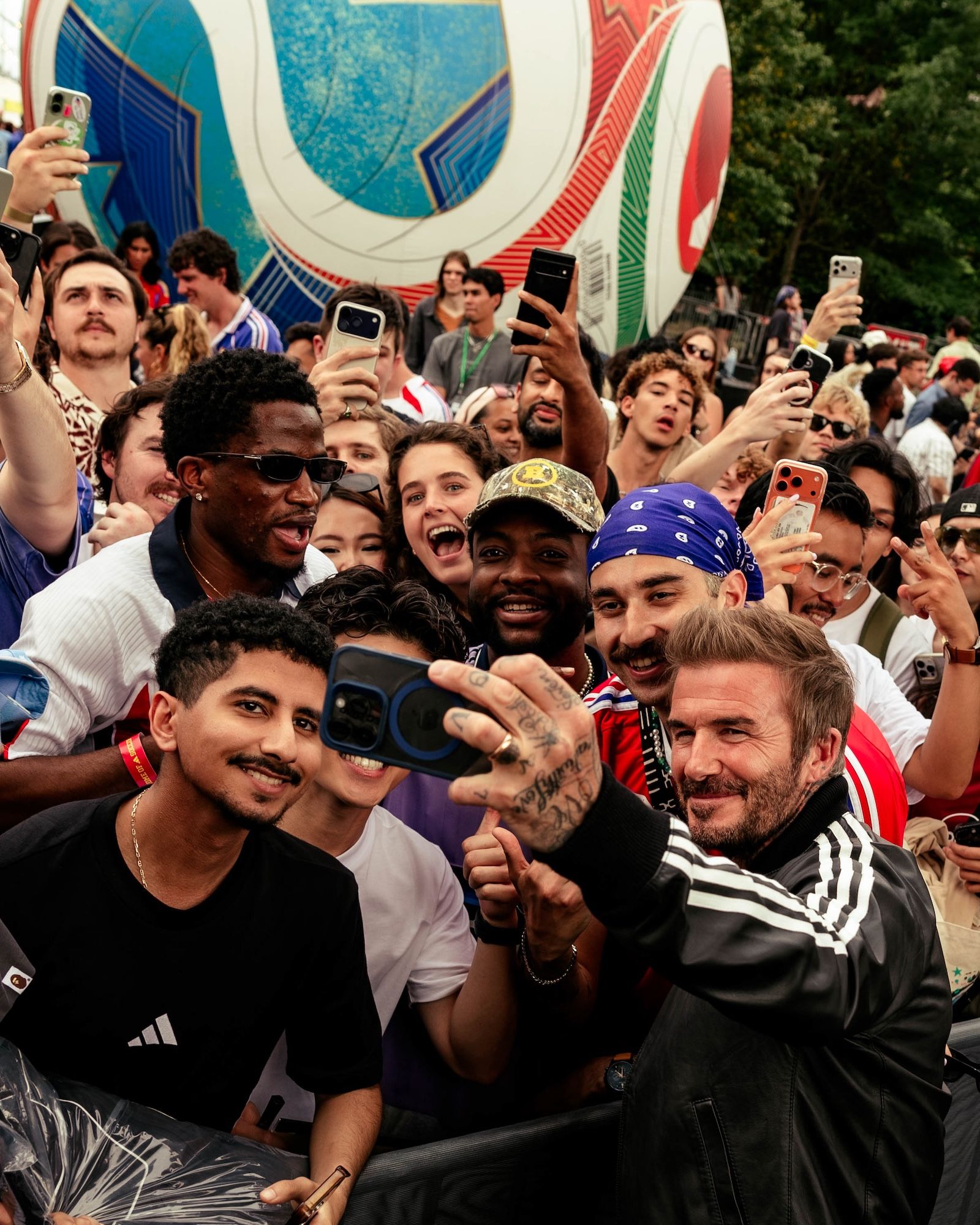 Crowd of people taking selfies and photos at an event, with a large colorful graphic backdrop visible.