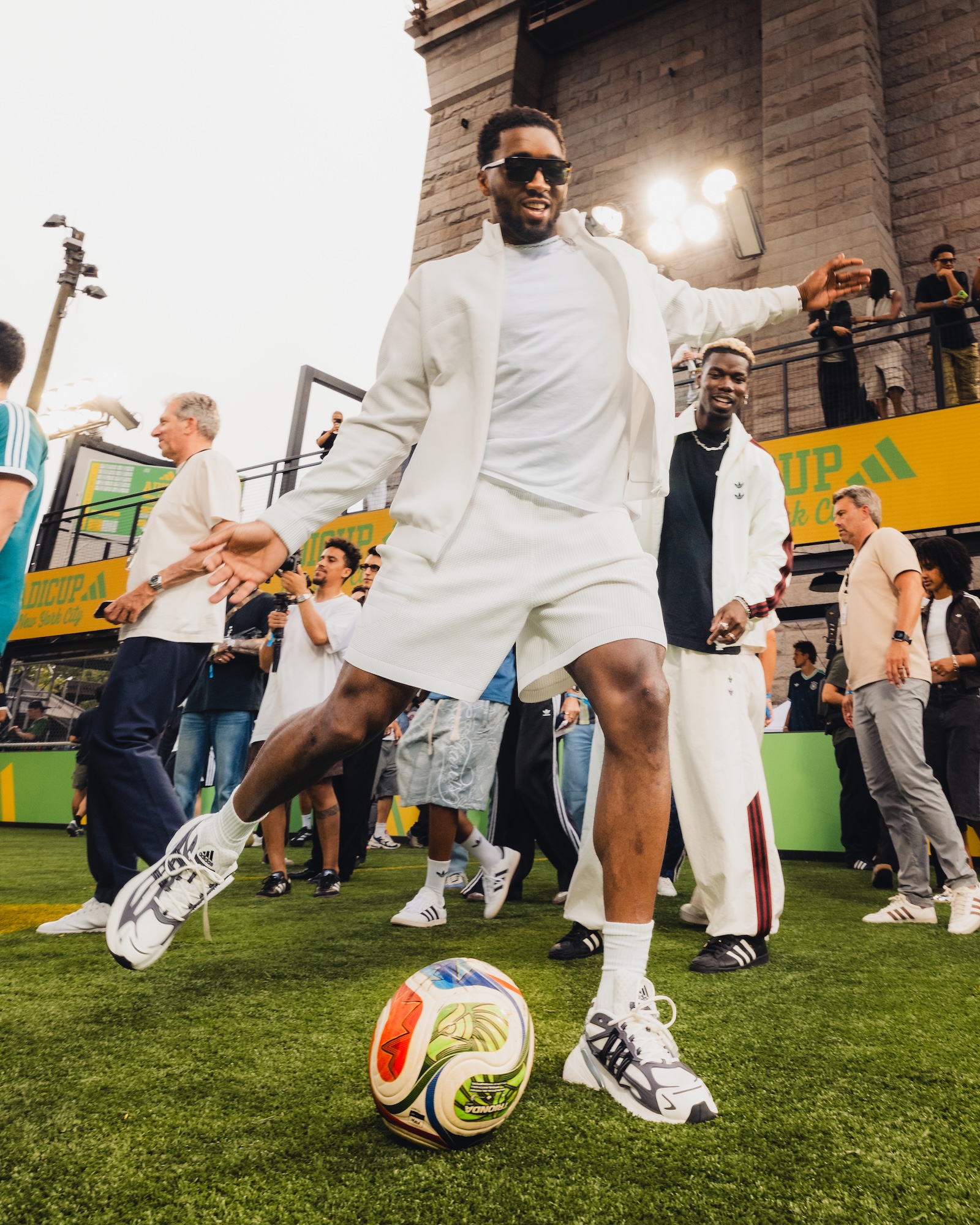A man in a white outfit and sunglasses standing on a soccer field, surrounded by a crowd and holding a soccer ball.
