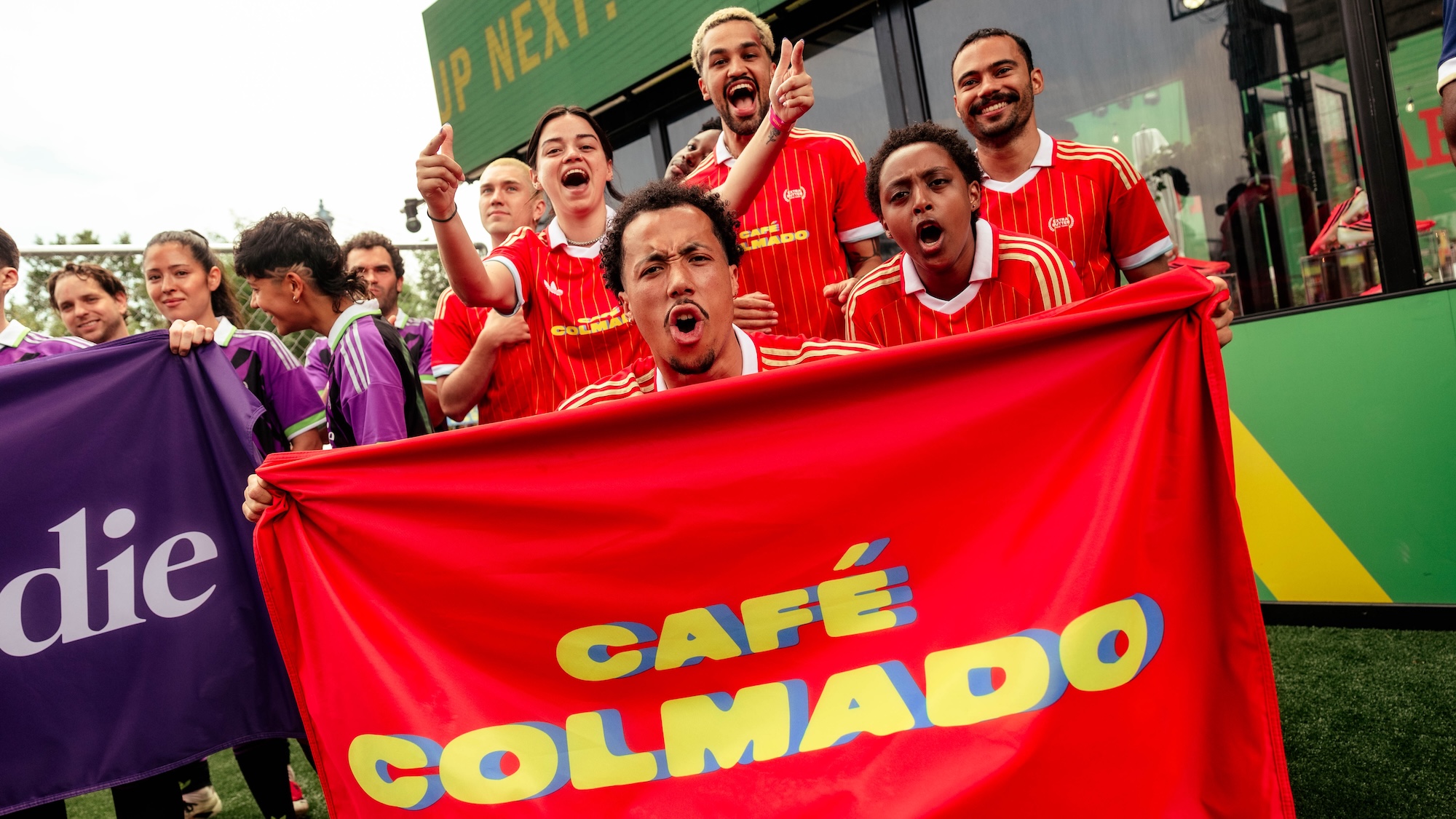 A group of people wearing red soccer jerseys cheering and holding a large red flag that says "CAFE GOLMADO".