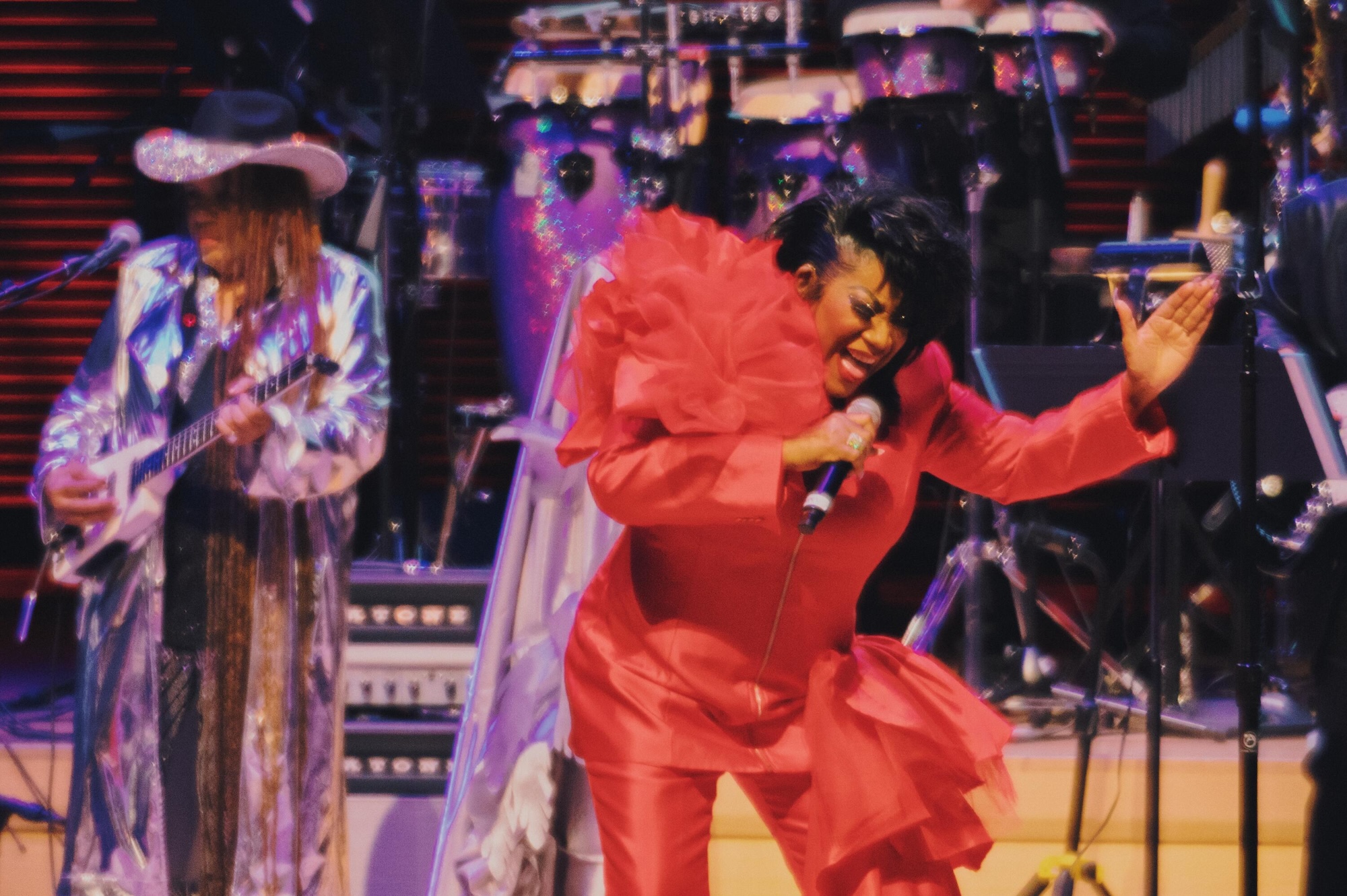 Two performers on stage, one wearing a red floral dress and the other in a colorful patterned outfit, singing and dancing energetically.