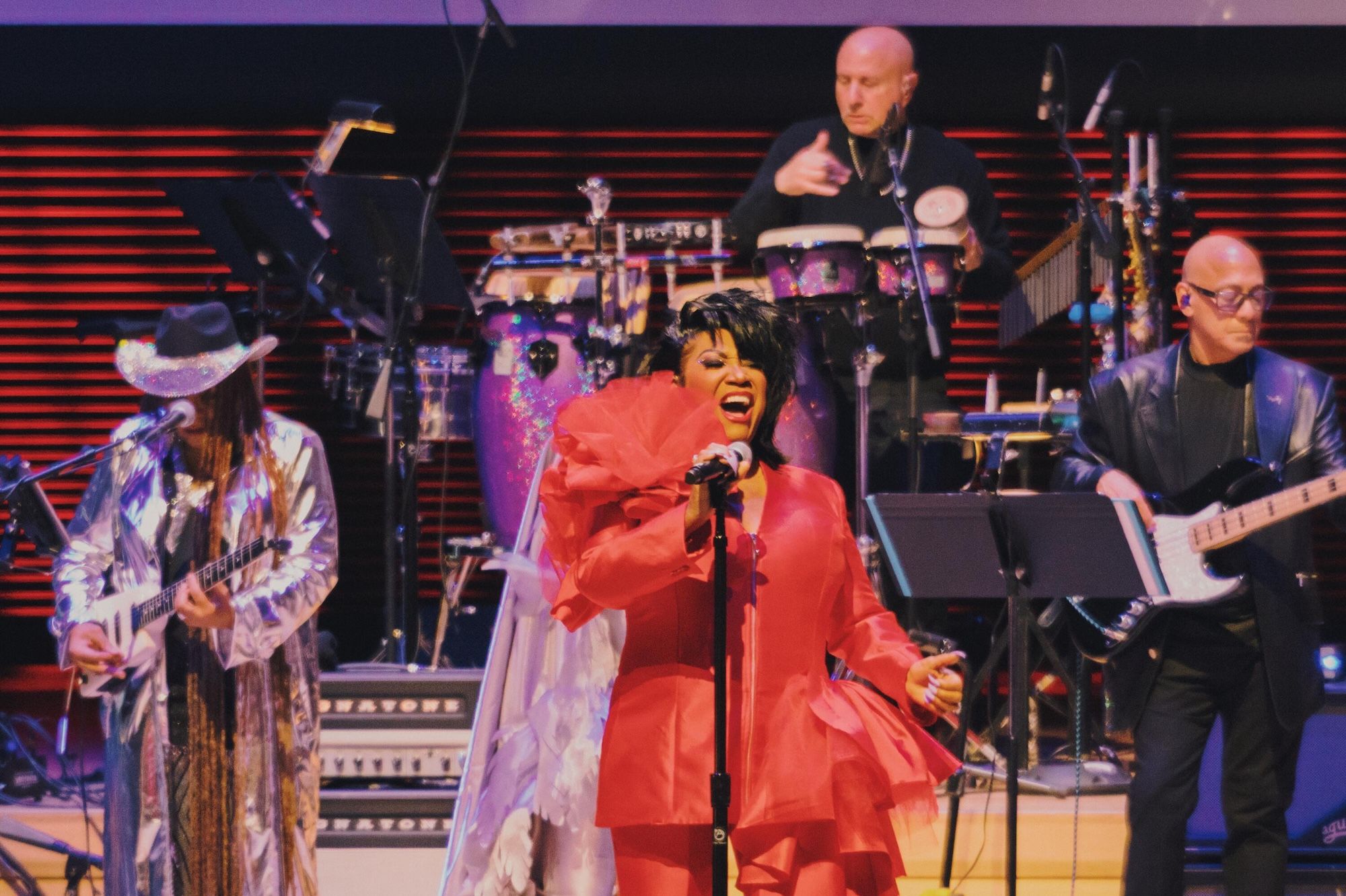 A performer in a red dress singing on stage, surrounded by musicians playing instruments.
