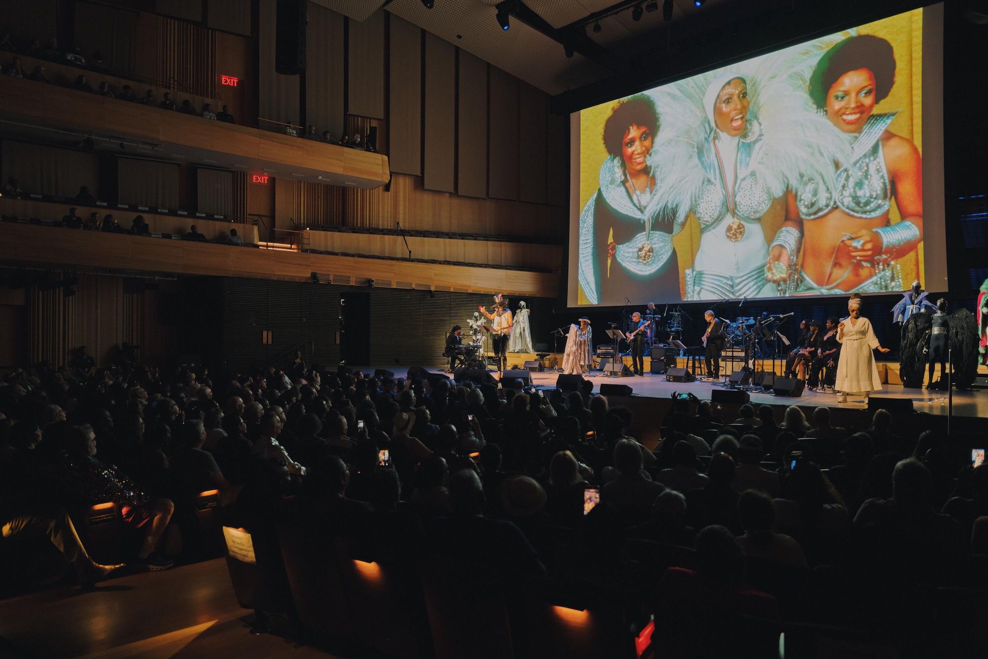 A large audience in a theater watching a performance on a large screen, which displays a colorful image of three women in elaborate costumes.