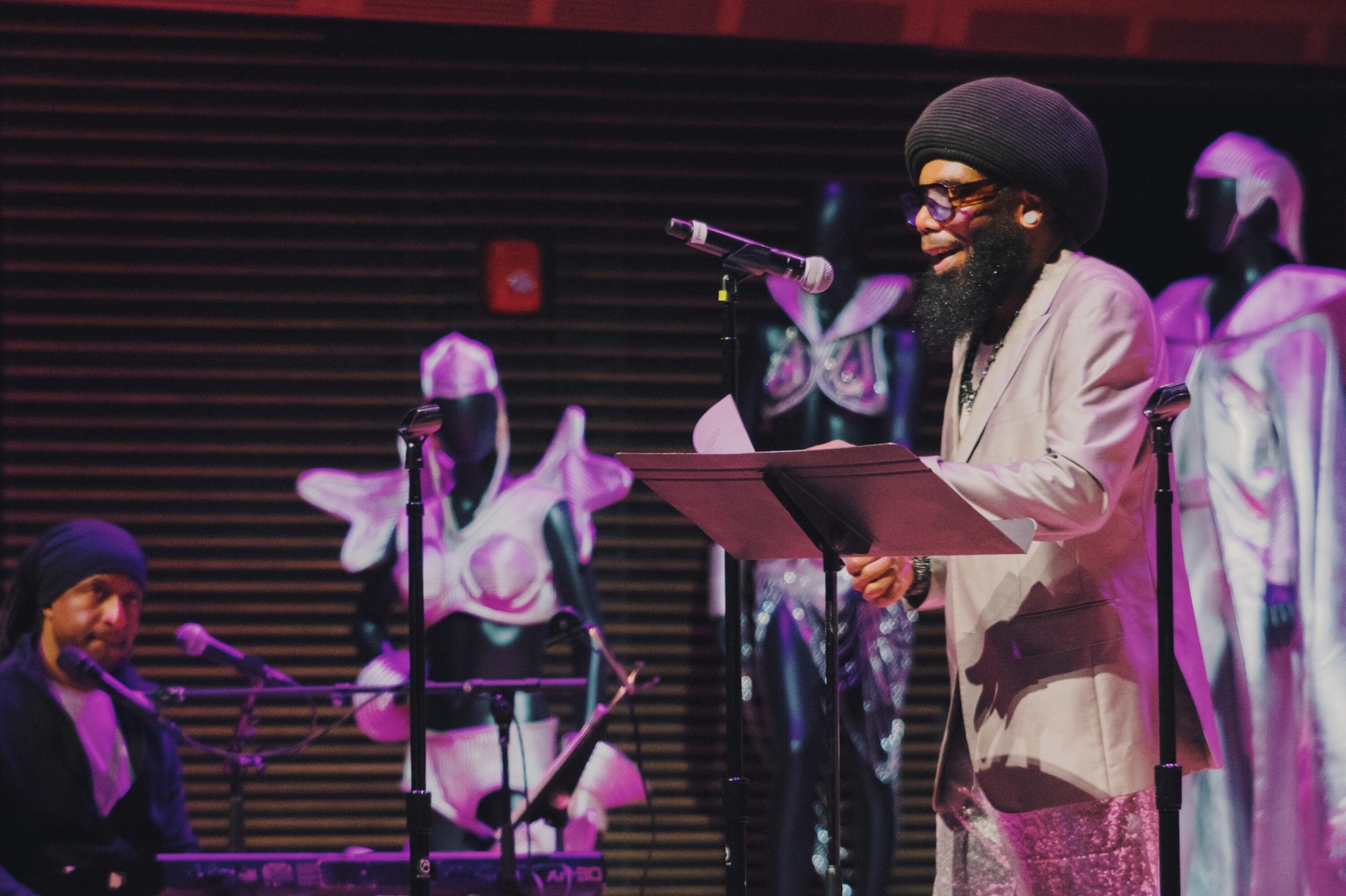 A performer wearing a white robe and turban stands at a microphone, reading from a book, surrounded by musicians on a dimly lit stage.
