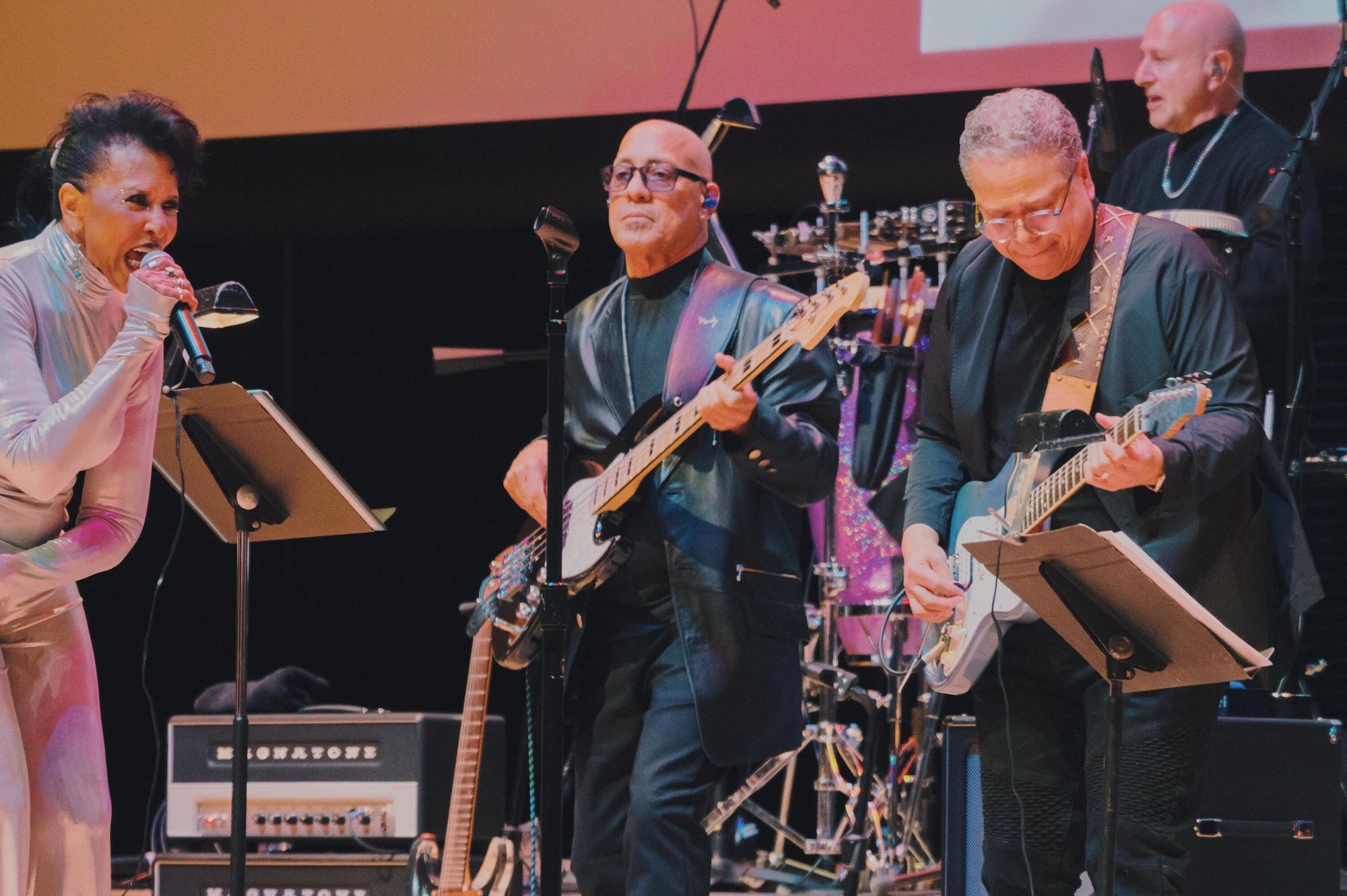 A group of older musicians performing on stage, including a woman singing into a microphone and two men playing electric guitars.