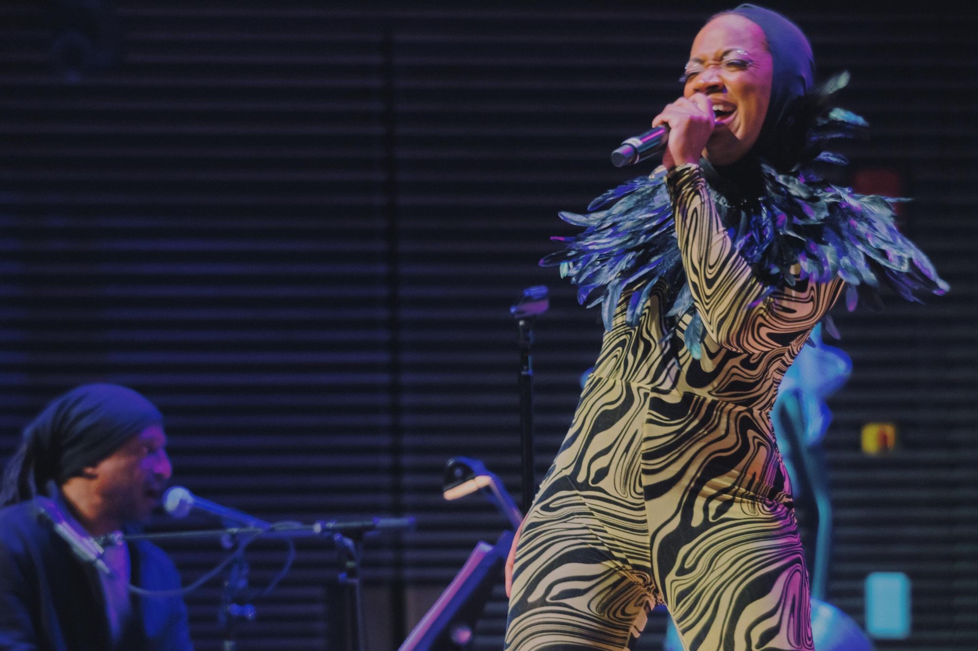 A performer in a colorful feathered costume singing into a microphone on stage, with a musician playing keyboard in the background.