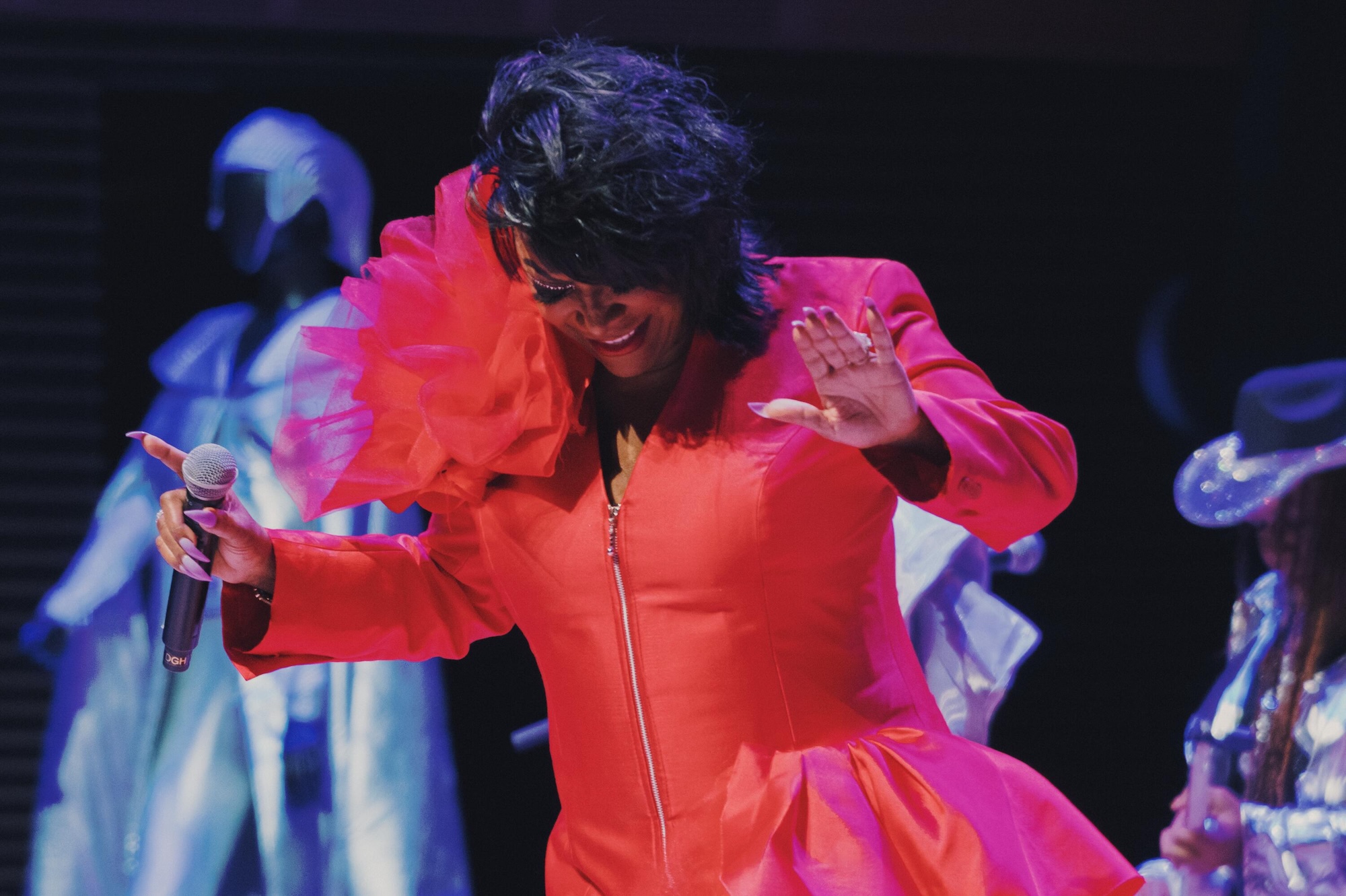 A person wearing a bright red dress with a large flower on the shoulder, dancing and singing into a microphone on a stage with colorful lighting.