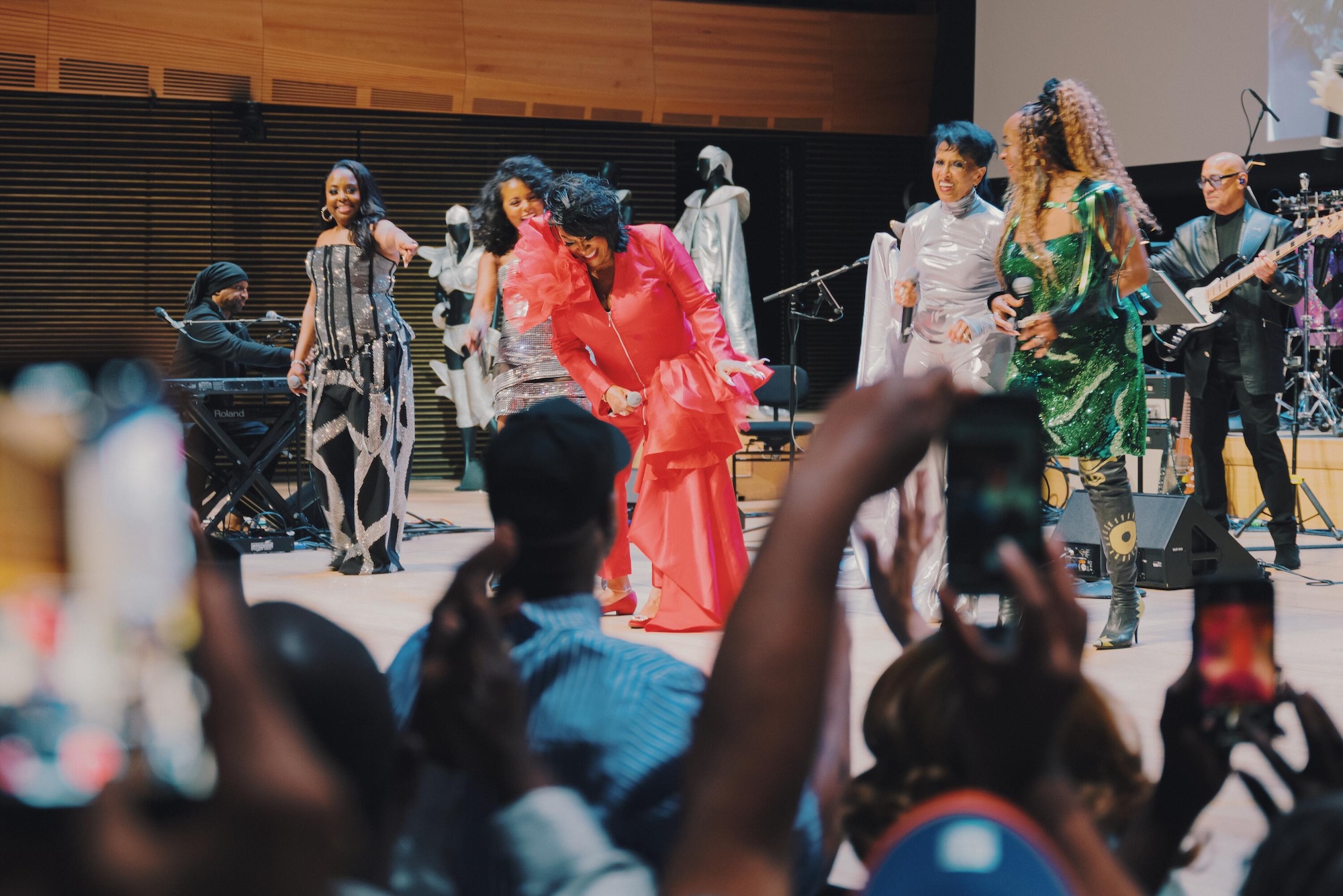 A group of musicians and dancers performing on stage, with the audience visible in the foreground. The performers are wearing colorful, elaborate costumes and appear to be in the midst of an energetic musical performance.