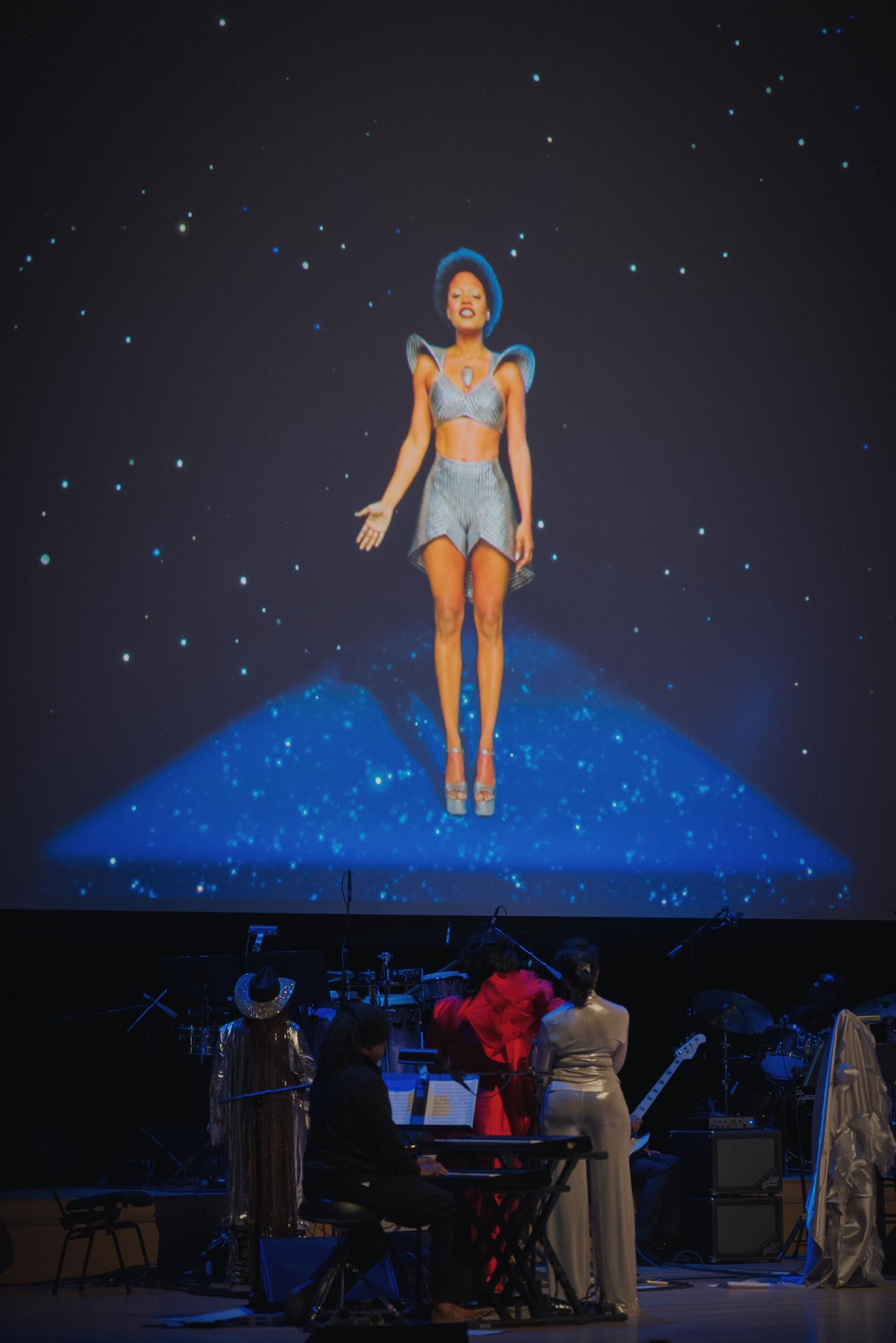 A holographic figure of a woman in a silver outfit appears to be performing on a stage, with a band and audience visible below.