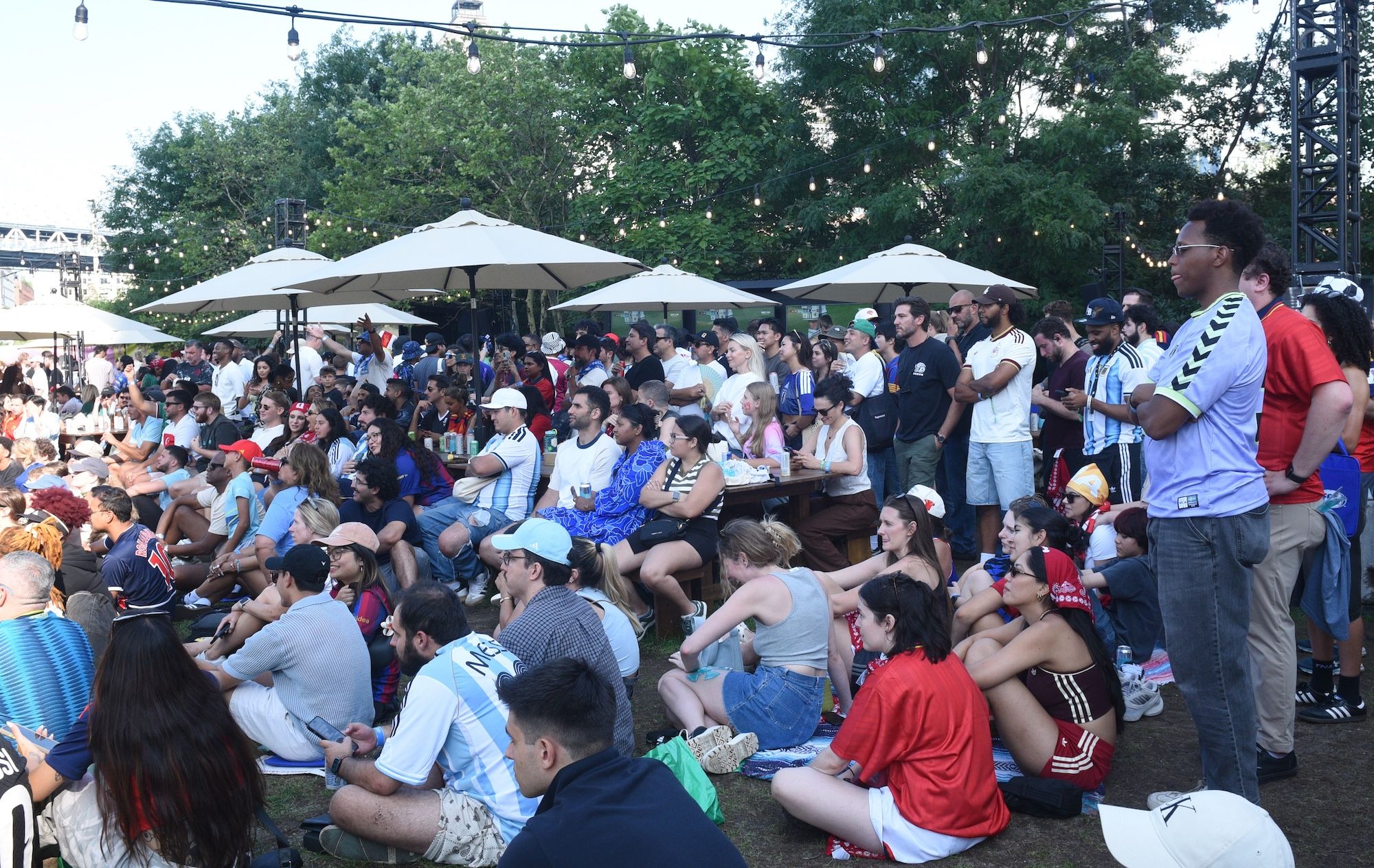 Crowded outdoor gathering with people sitting on the ground and standing under string lights and umbrellas.