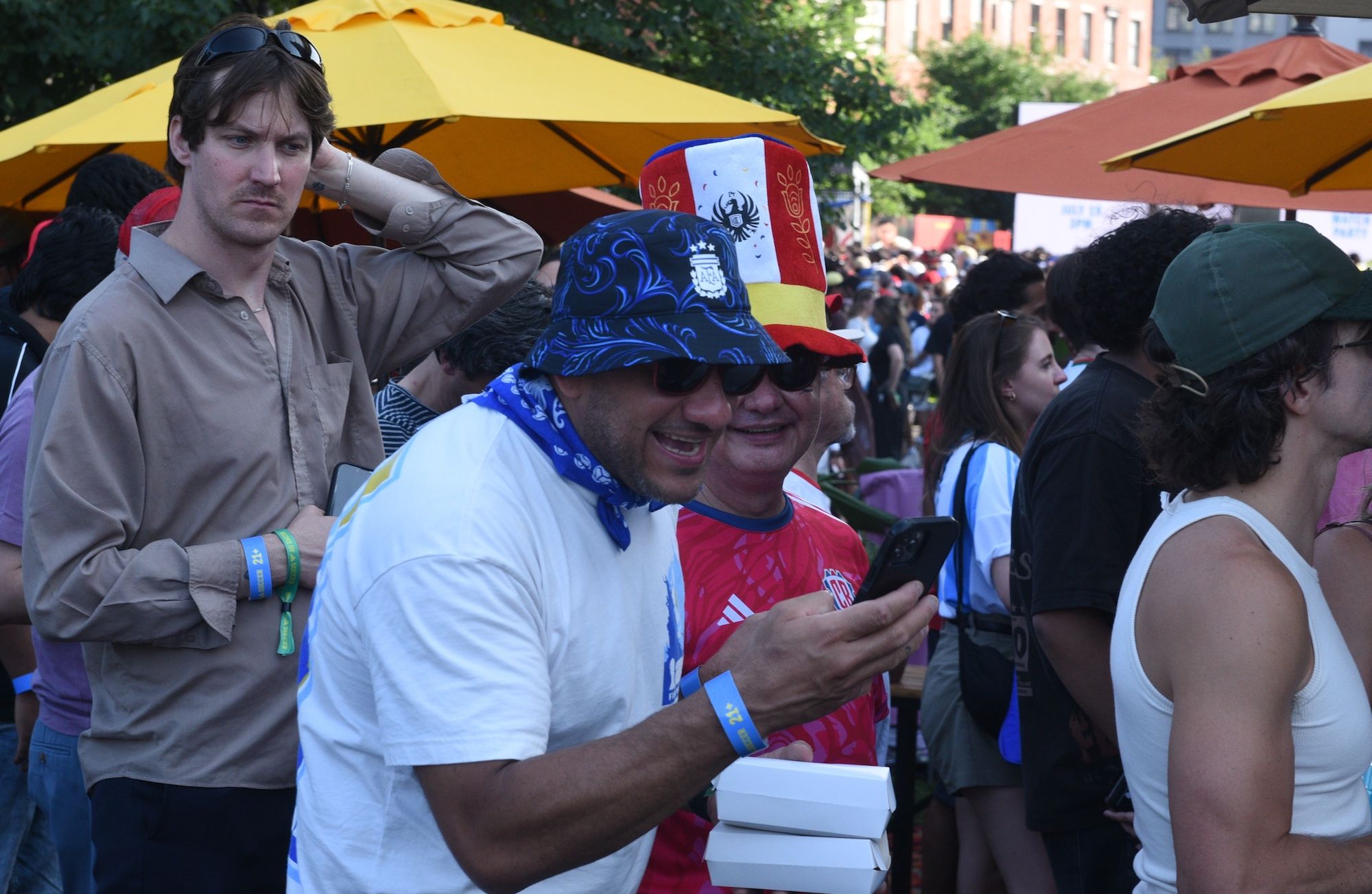 A crowd of people at an outdoor event, with a man wearing a blue and white hat and a red shirt standing in the foreground holding a mobile device.