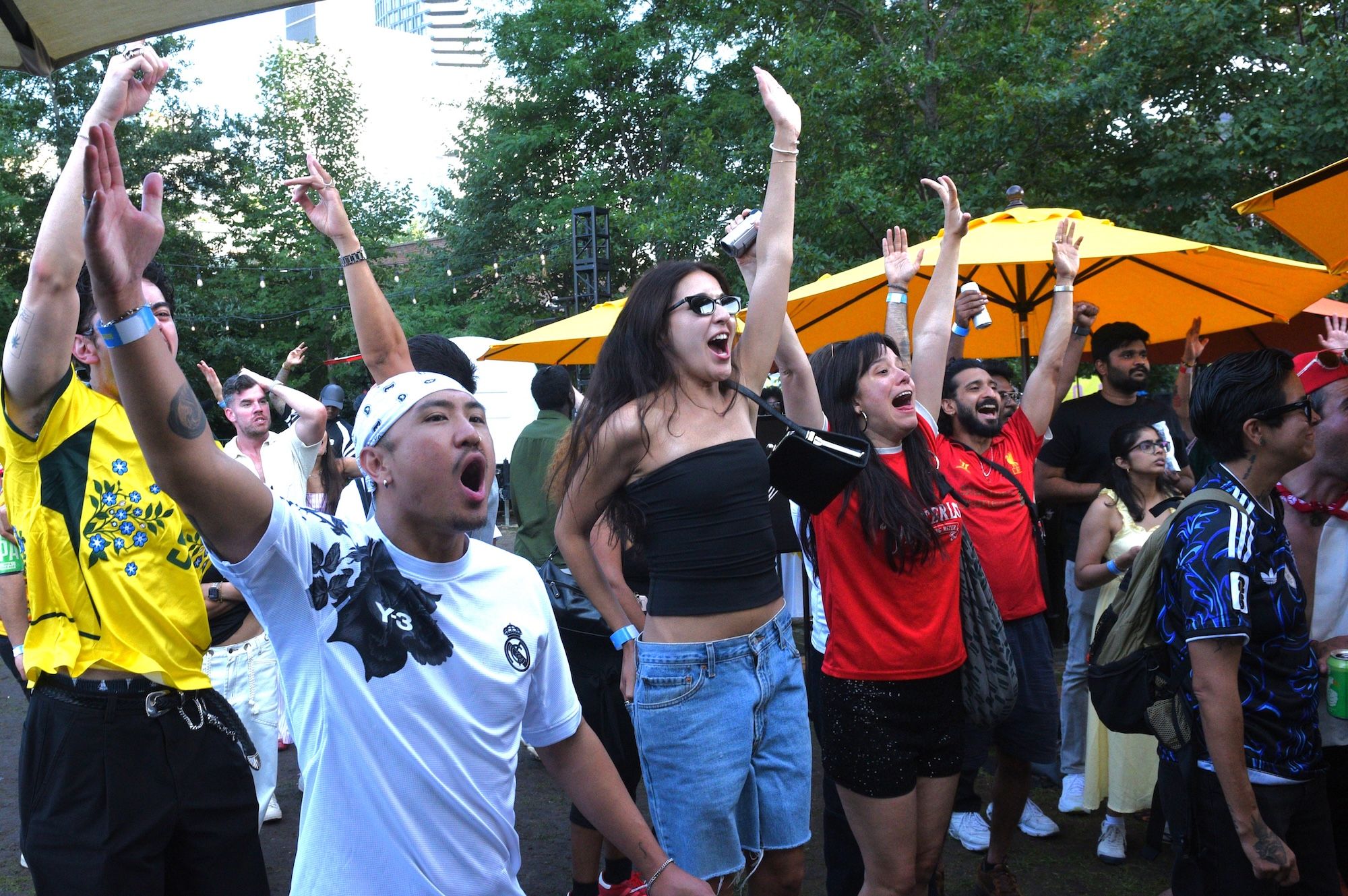 A crowd of people cheering and raising their arms at an outdoor event, with orange umbrellas and trees in the background.