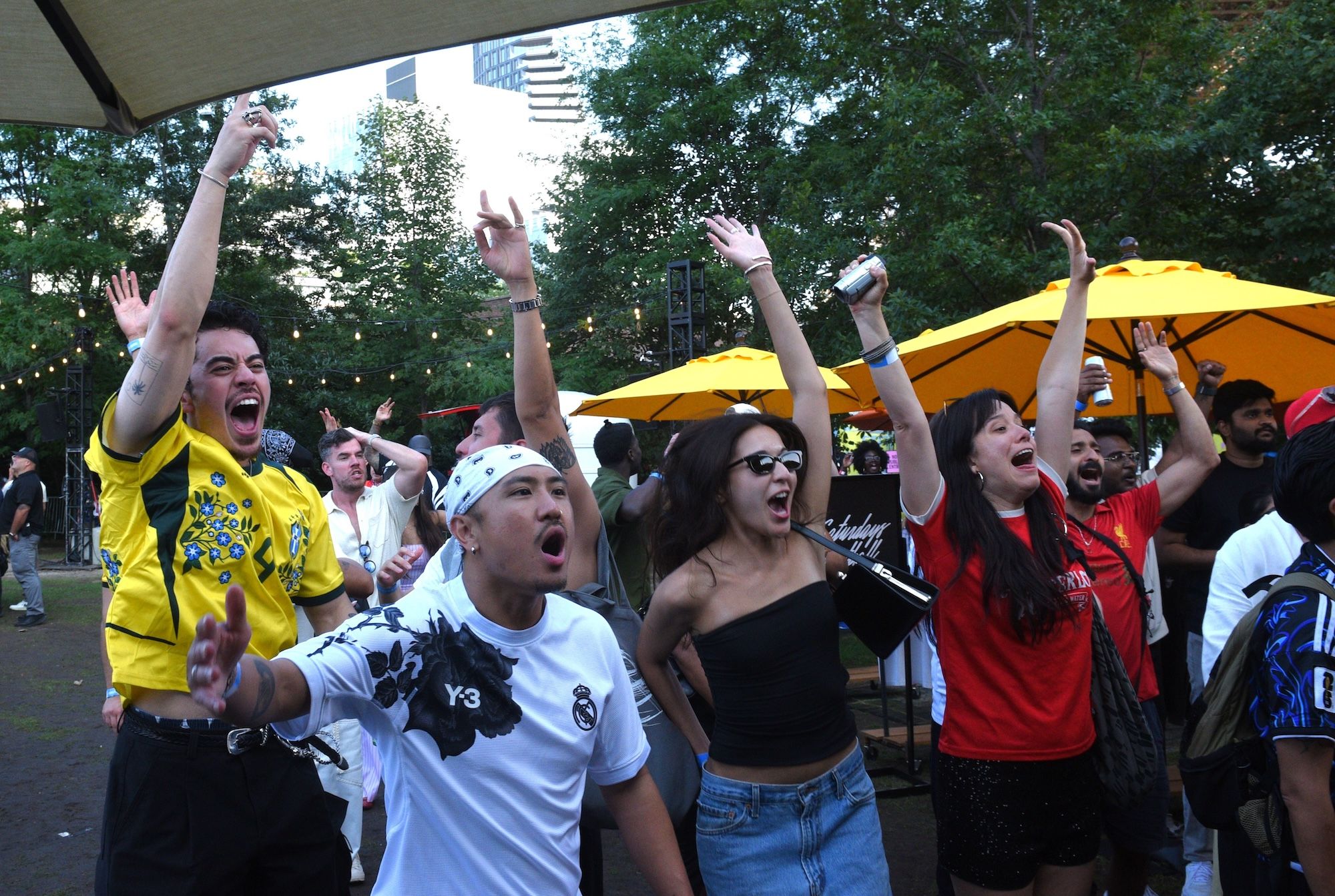 A crowd of people cheering and raising their arms at an outdoor event, with yellow umbrellas and a large building in the background.