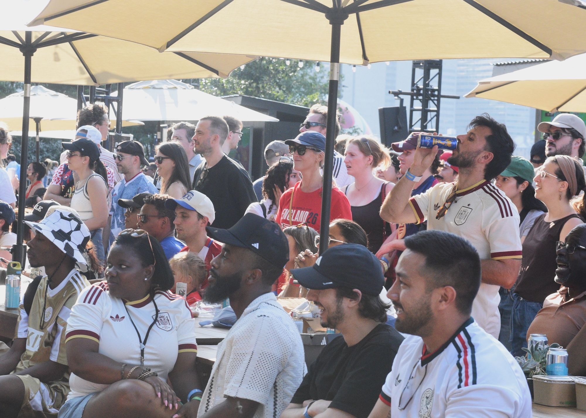 Crowded outdoor dining area with people wearing sports jerseys and casual clothing, umbrellas, and a stage or performance area visible in the background.