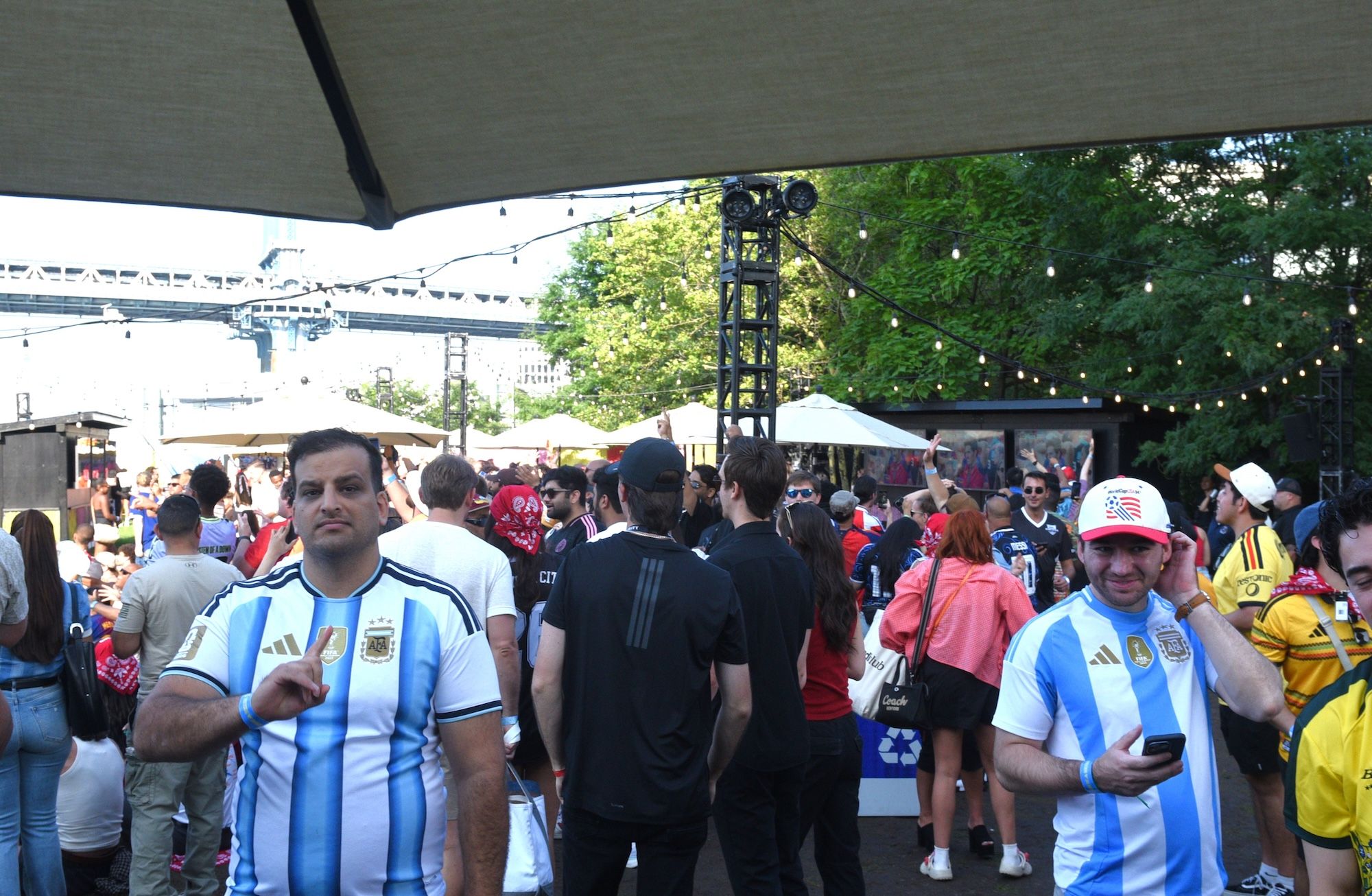 Crowded outdoor event with people wearing Argentina and USA soccer jerseys, surrounded by tents, string lights, and a tall camera tower.