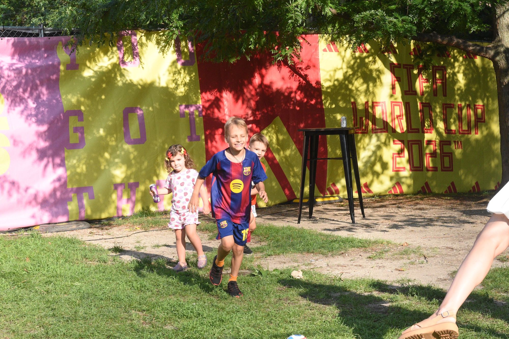 Two children playing outdoors in a colorful, graffiti-covered environment with a Barcelona FC jersey and a stool.