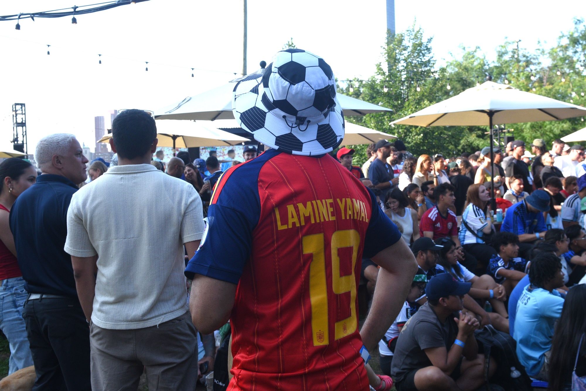 A crowd of people at an outdoor event, with a person wearing a soccer jersey with the name "LAMINE YAMAR 19" on the back and a large soccer ball-shaped hat.