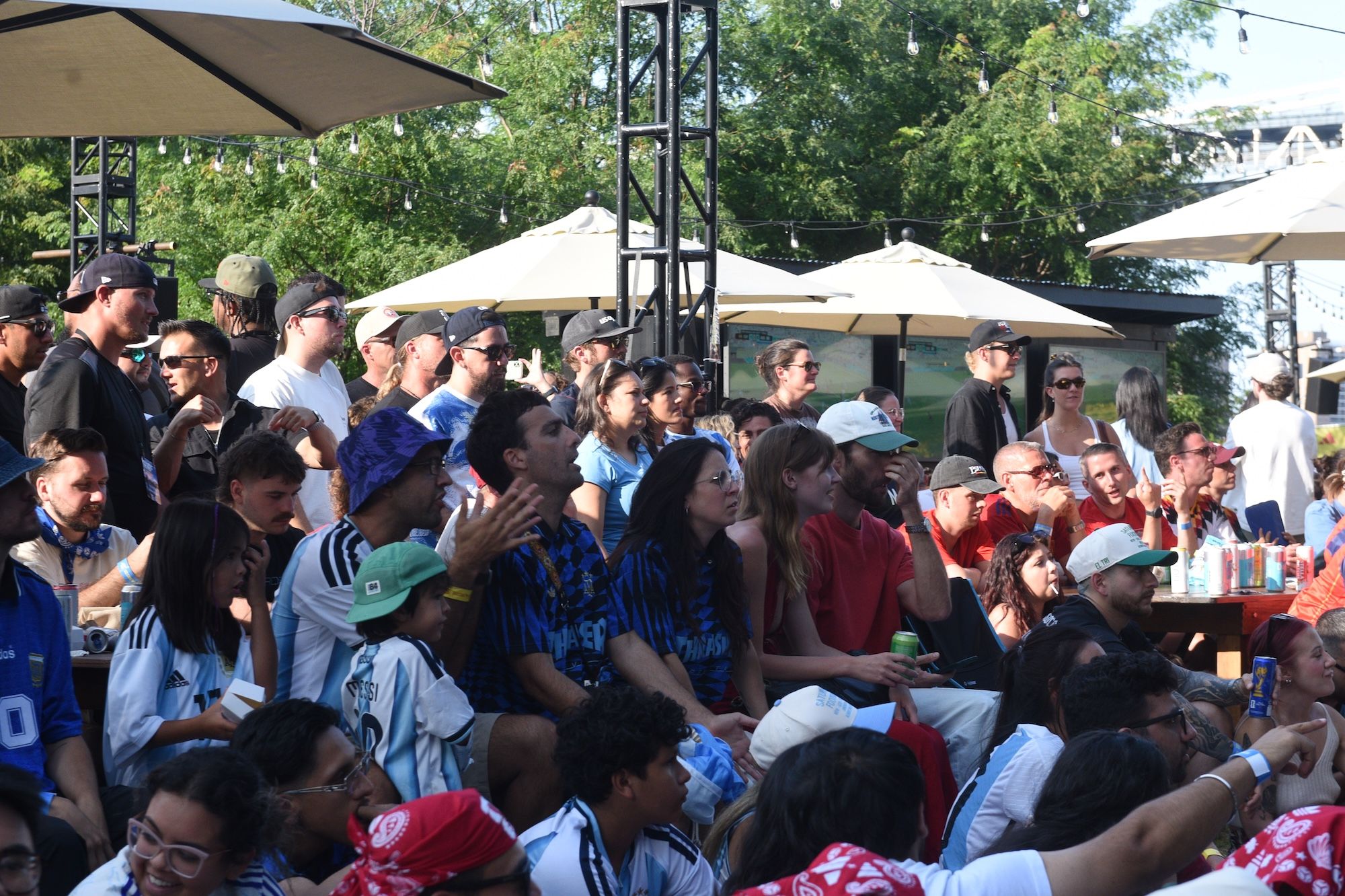 Crowded outdoor event with people sitting at tables under umbrellas, surrounded by trees.