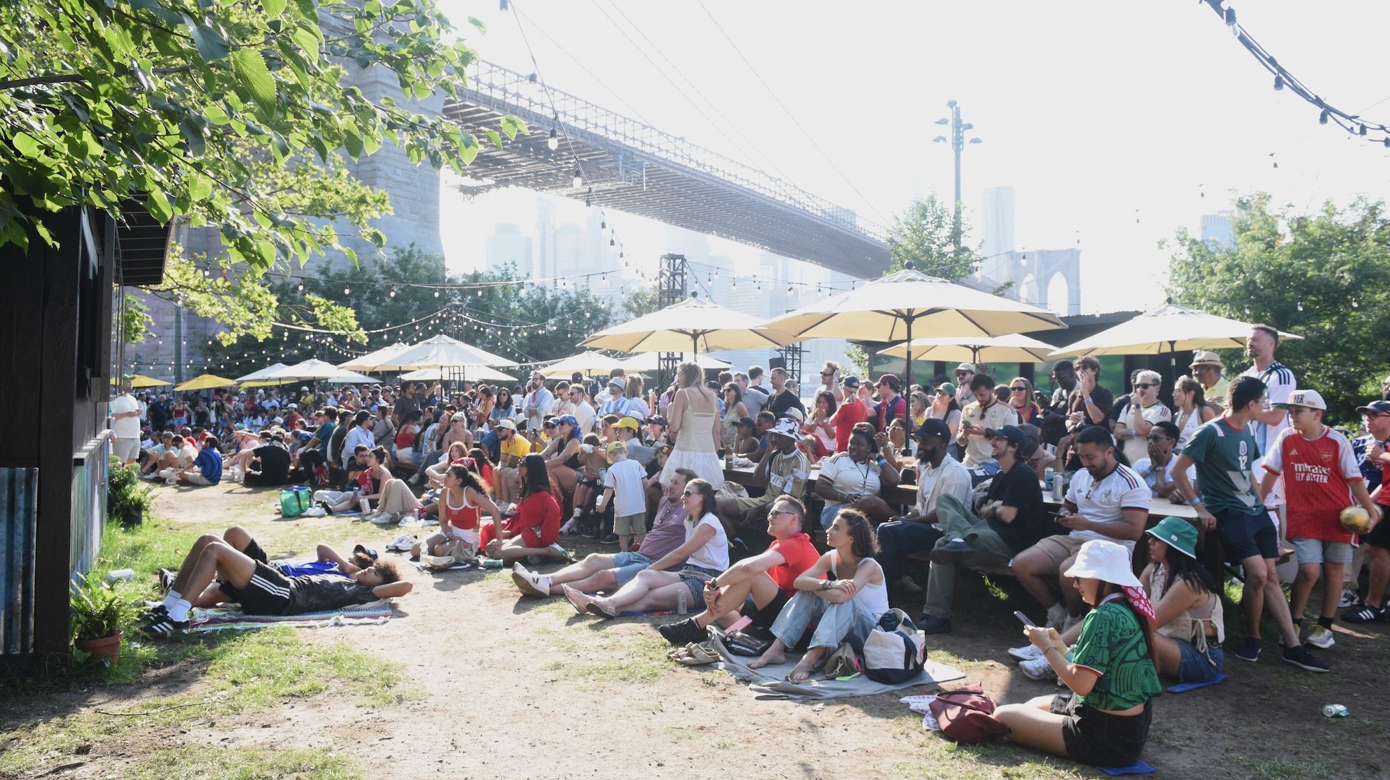 Crowded outdoor event with people sitting on the ground under umbrellas, with a large bridge visible in the background.