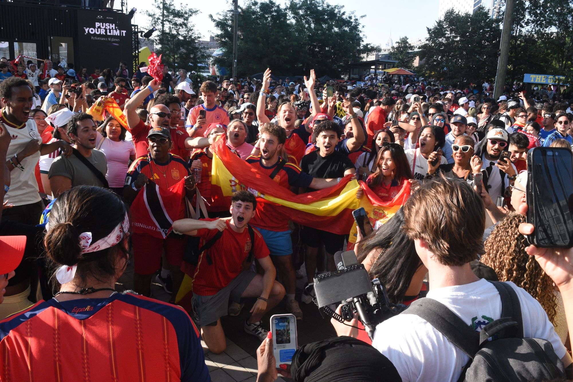 Crowd of people in red shirts waving Spanish flags, cheering and celebrating at an outdoor event or rally.