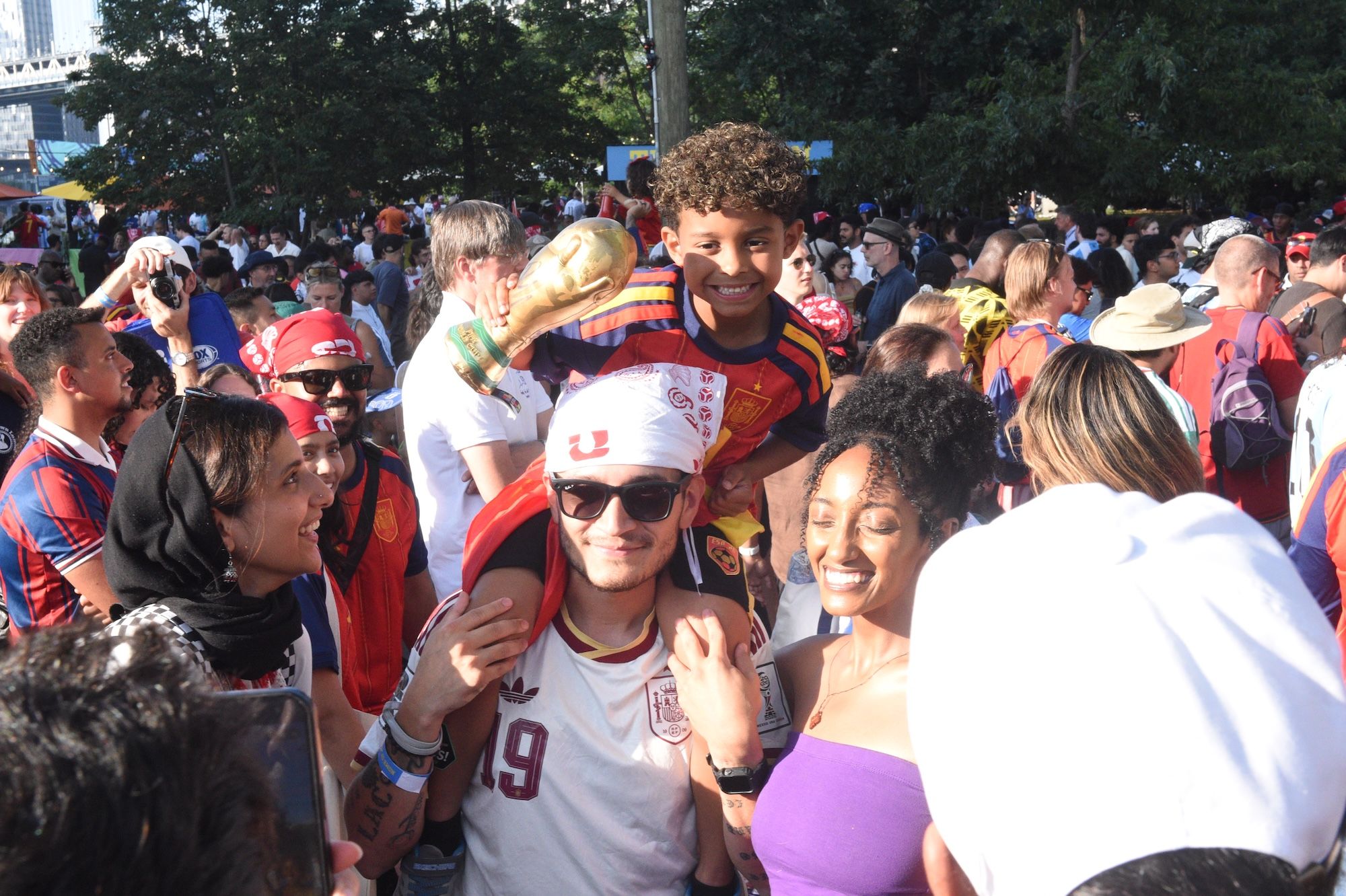 Crowd of people in sports jerseys and costumes celebrating at an outdoor event, with a smiling young person in the foreground.