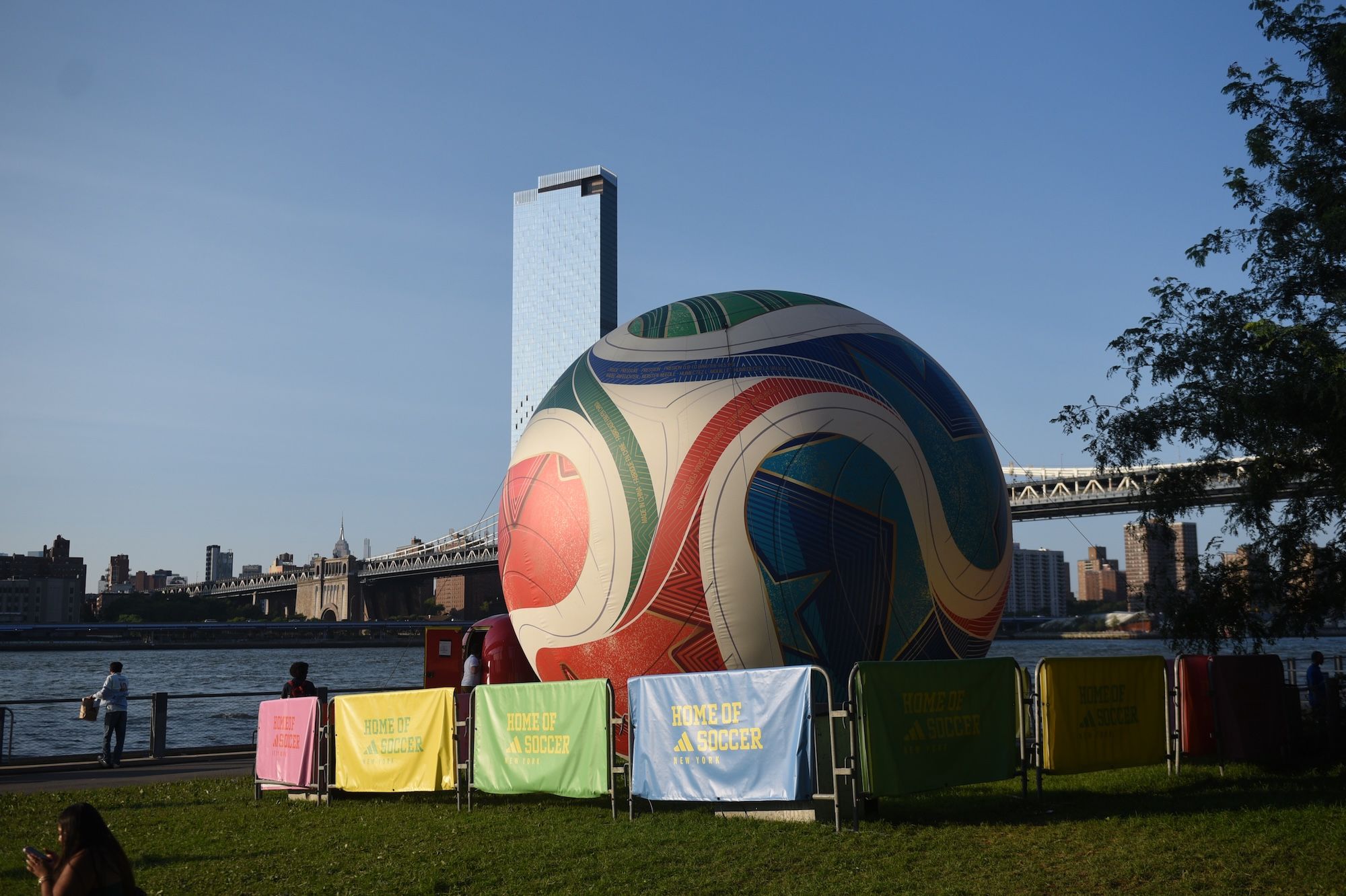 A large inflatable soccer ball sculpture on a grassy field overlooking the East River and Manhattan skyline, with banners reading "Home of Soccer" displayed nearby.